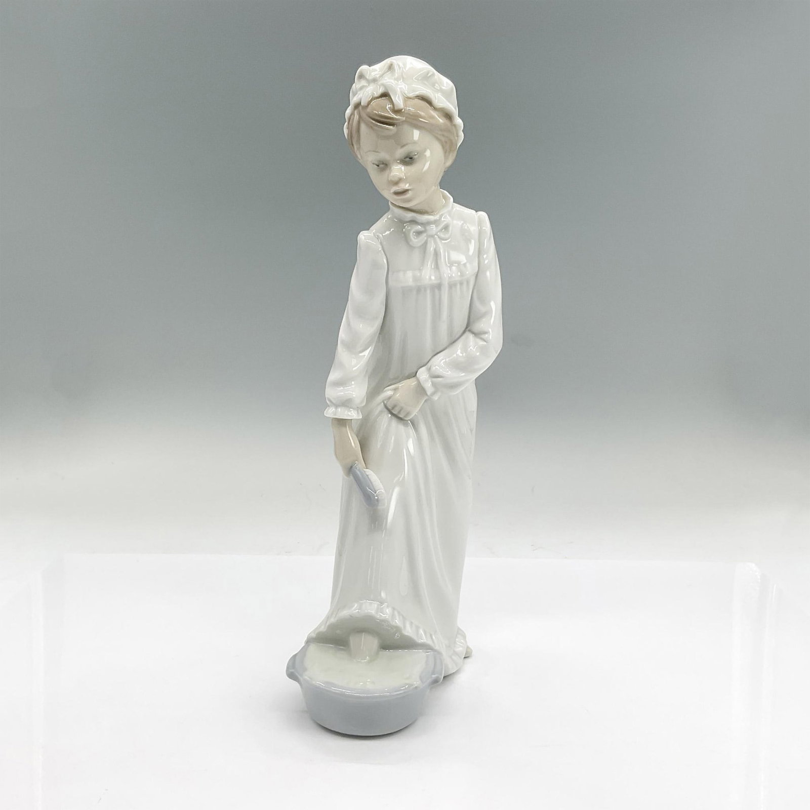 Nao By Lladro Porcelain Figurine, Girl In Nightgown: Glazed porcelain figure of a girl in a nightgown and nightcap getting ready for bed, washing her feet. Nao backstamp. Dimensions: 5"L x 3"W x 11"H Manufacturer: Nao by Lladro Country of Origin: Spain