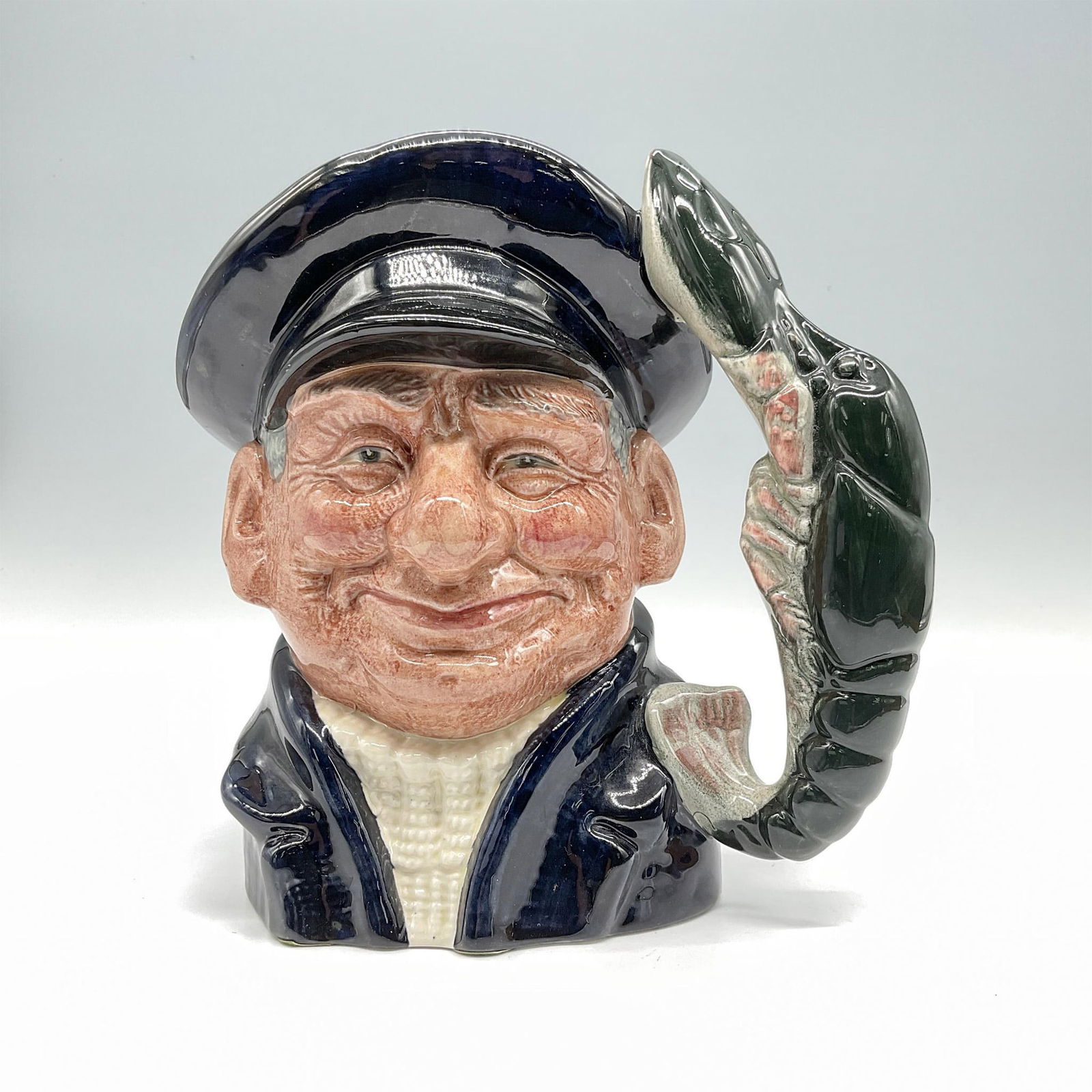 Lobster Man D6617 - Large - Royal Doulton Character Jug: Features a dark blue jacket and cap paired with a white fisherman's jersey, creating a classic and nautical - inspired ensemble. Royal Doulton backstamp. Artist: David B. Biggs Issued: 1968-1991 Dimen