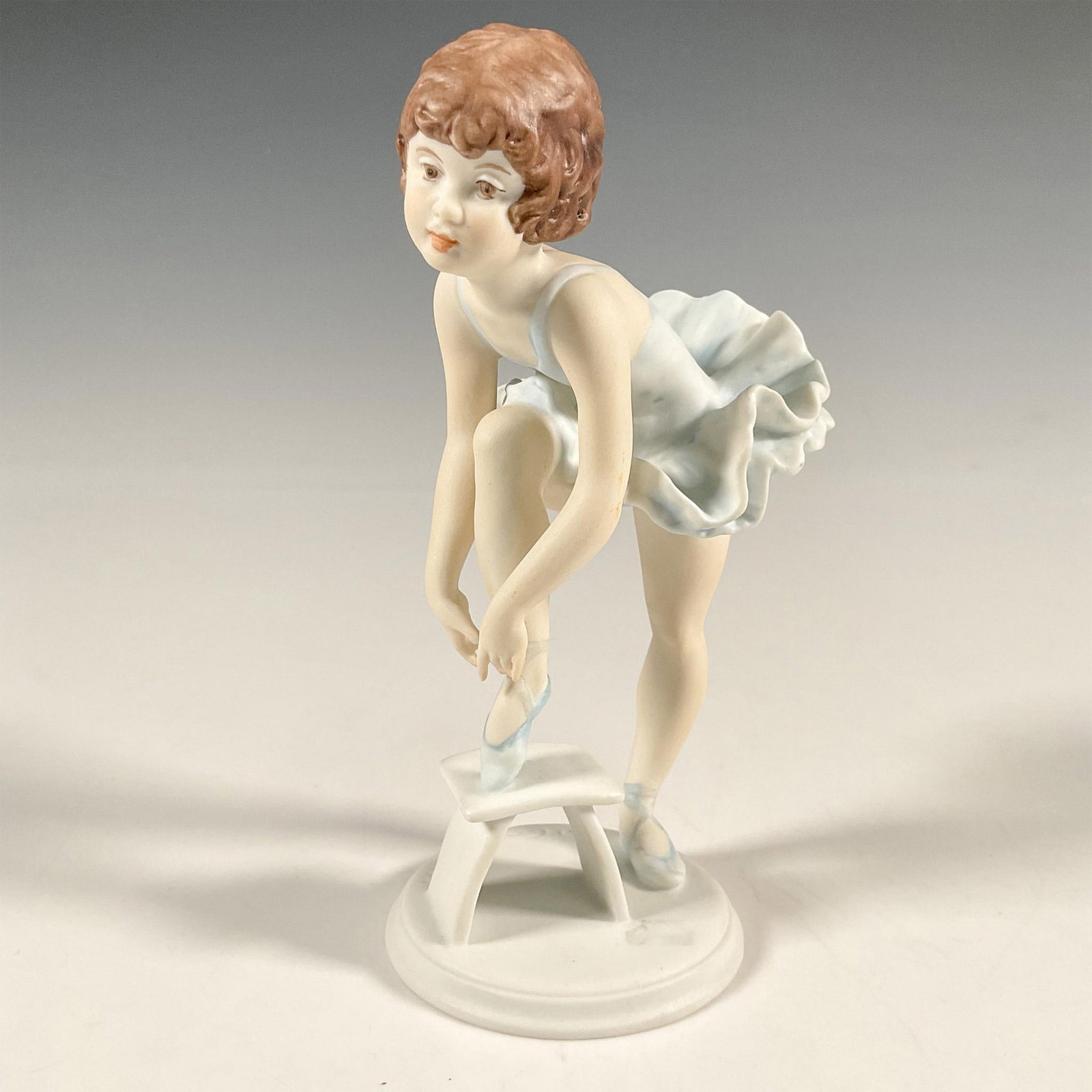 Kaiser Porcelain Figurine, Ballerina: A colored bisque porcelain figure modeled as a young ballerina posing as she laces her pointe shoe. Kaiser Signed by artist to base. Germany backstamp. Artist: Wolfgang Gawantka Dimensions: 3"L x 2.75