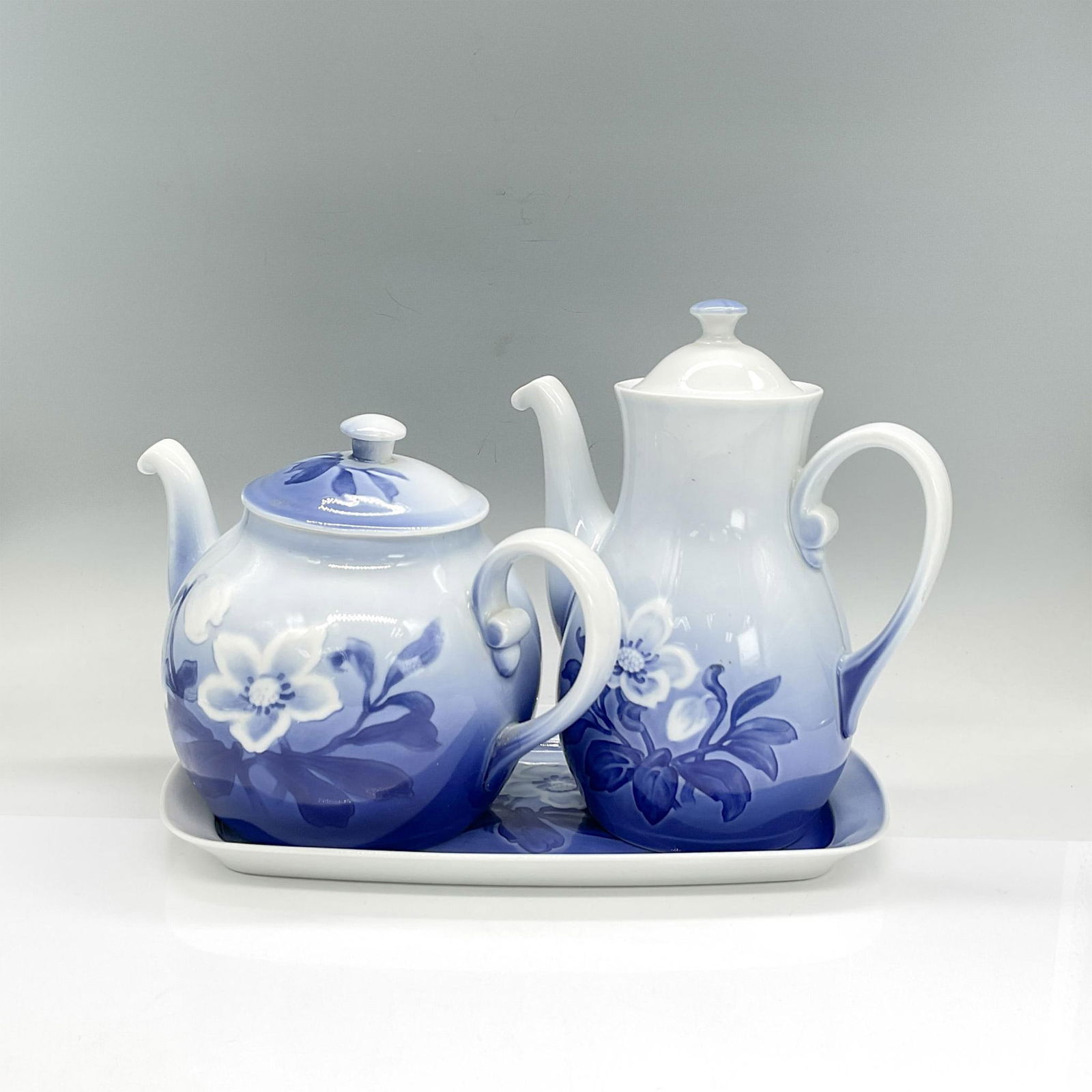 3pc Bing & Grondahl Teapot/Coffee Pot/Tray, Christmas Rose: Porcelain serveware in a design of a blue background with white Christmas Roses. Teapot 9"L x 5"W x 6.25"H. Coffee pot 7.25"L x 3.25"W x 8"H. Appetizer platter 11"L x 6"W x 0.75"H. Bing & Grondahl bac