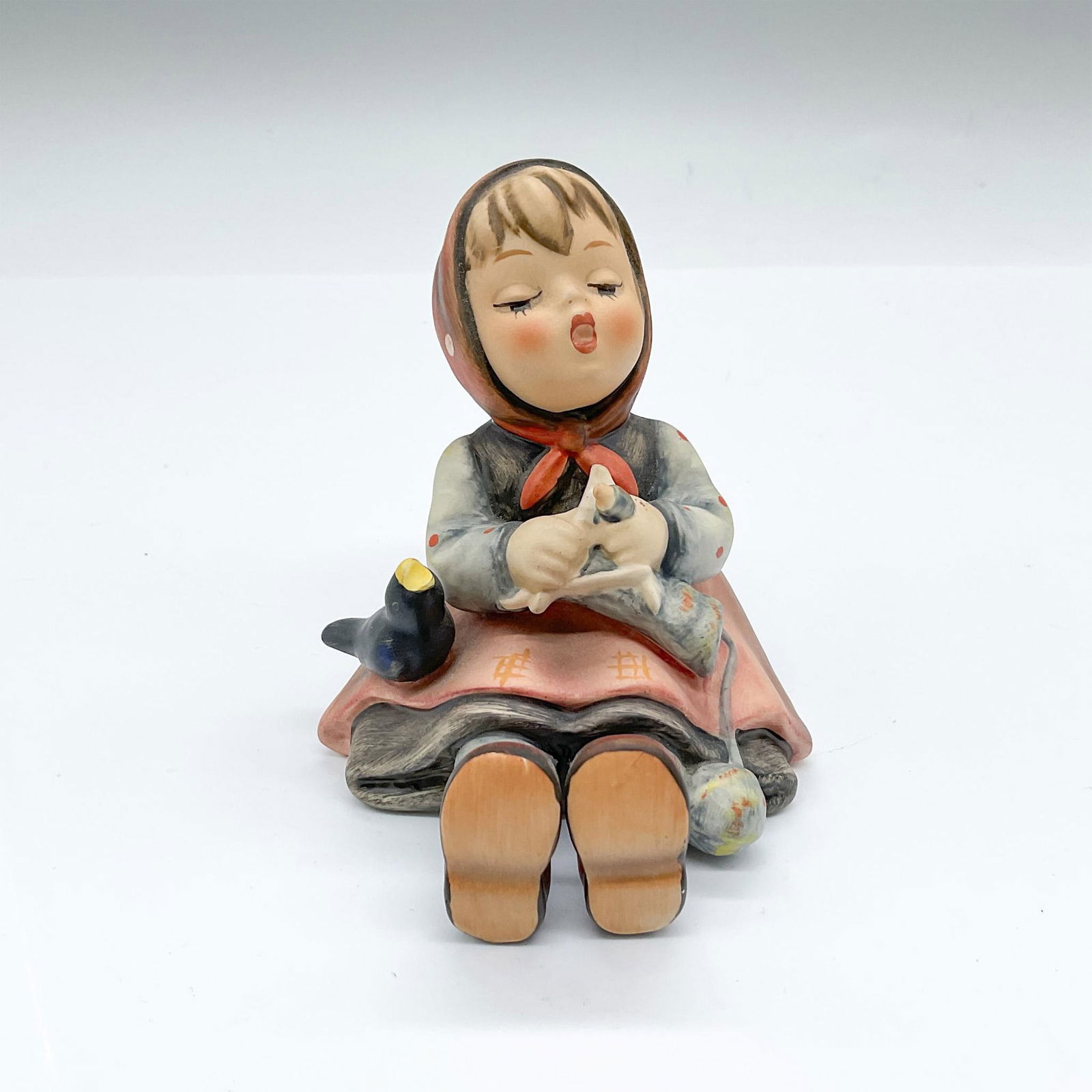 Goebel Hummel Porcelain Figurine, Happy Pastime: Hand painted figure of a seated girl knitting while she is singing. Goebel backstamp. Issued: 20th century Dimensions: 3.5"L x 3"W x 3.5"H Manufacturer: Goebel Hummel Country of Origin: Germany