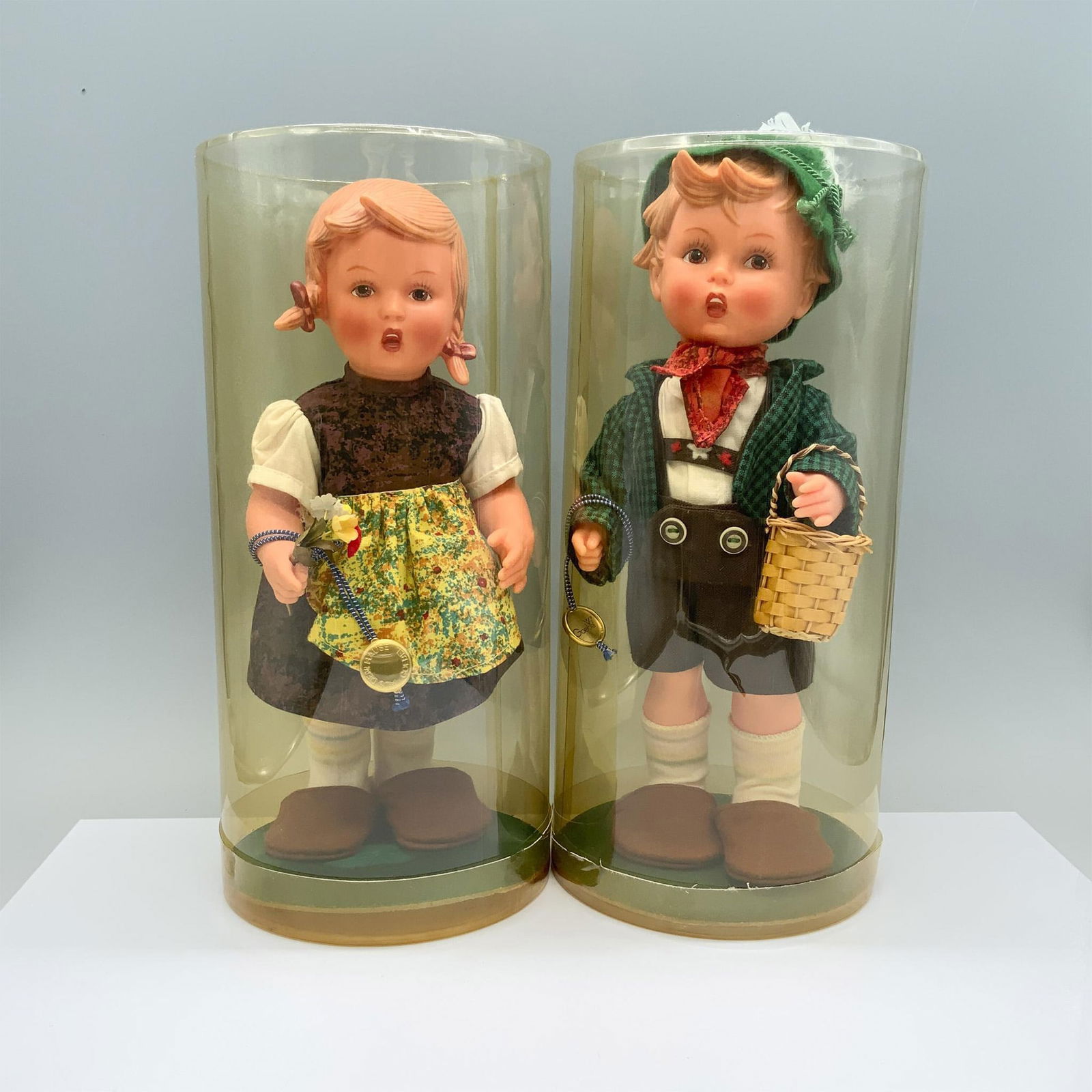 2pc Goebel Hummel Dolls, Happy Wanderers: In this charming the boy is dressed in a green jacket with brown overalls and carries a basket, while the girl wears a brown dress complemented by a yellow apron and holds a small bouquet of flowers.