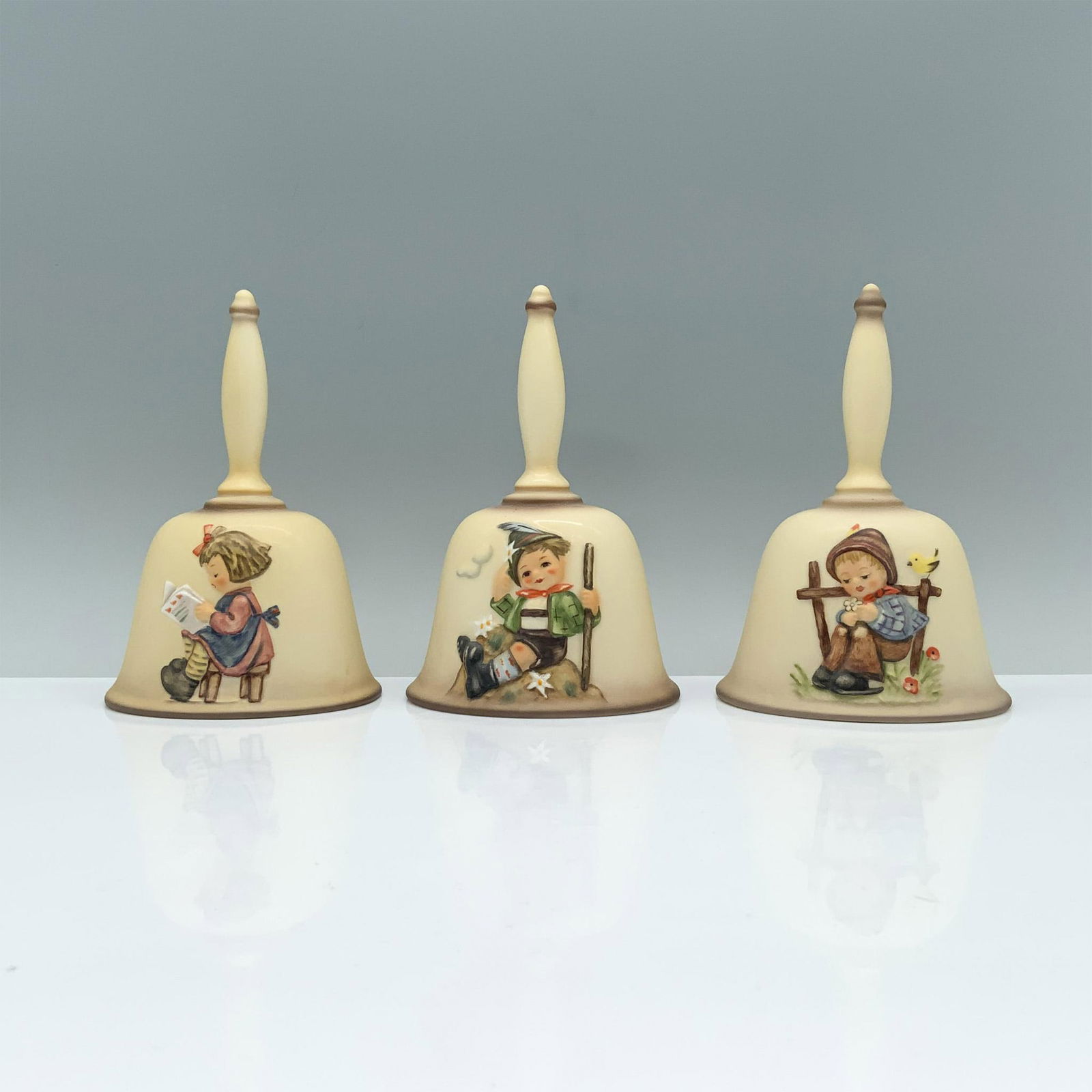 3pc Goebel Hummel Annual Bells, HUM 704, 706, 712: In this delightful set you'll find scenes of a boy admiring a flower with a canary by his side (1982), a boy sitting at the top of a dirt mound with his walking stick (1984), and a girl reading peacef