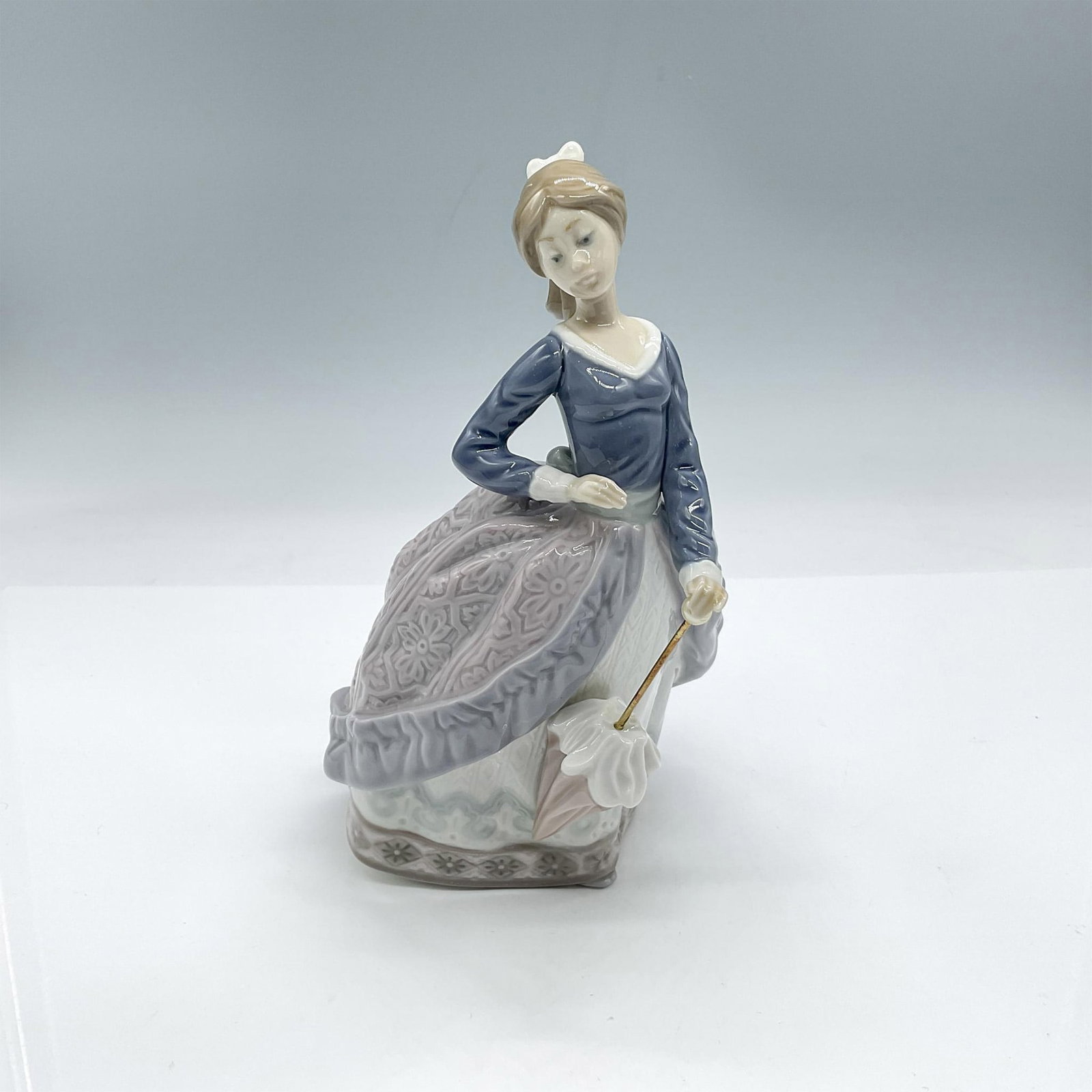 Evita 1005212 - Lladro Porcelain Figurine: Glossy, hand made porcelain. Depicts a girl leaning on an umbrella with pearl and metal handle, wears a textured dress in shades of purple and blue. Lladro backstamp. Artist: Jose Puche Issued: 1984 -