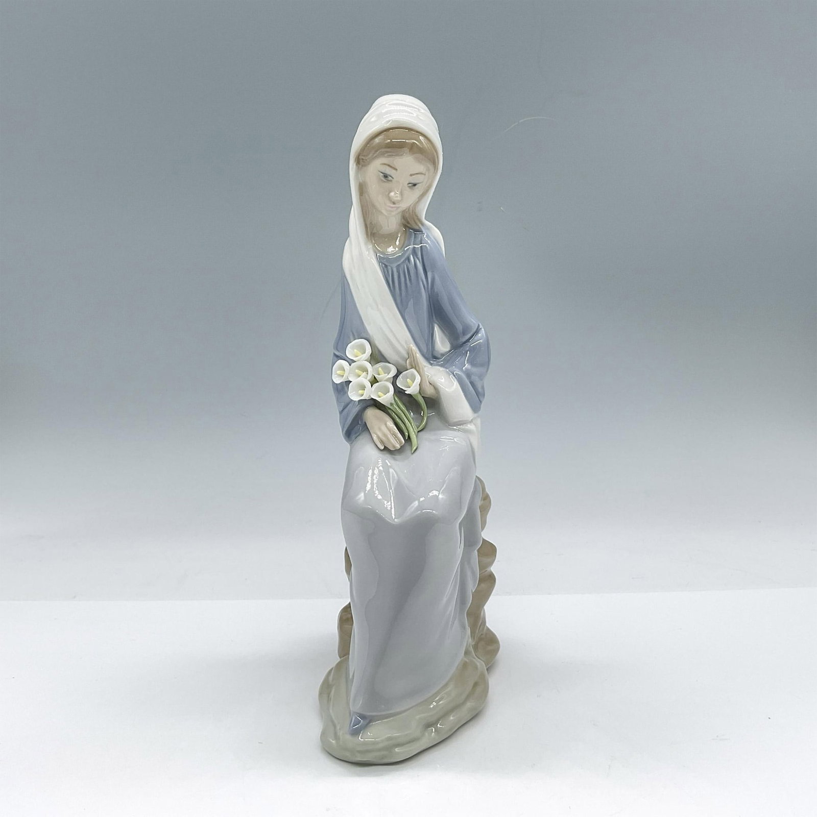 Girl with Lilies 1004972 - Lladro Porcelain Figurine: Glossy, hand made porcelain crafted in pale pastel colors, captures a serene scene. The girl dressed in a soft, pastel blue gown with a gentle drape, complemented by a white veil that frames the figur