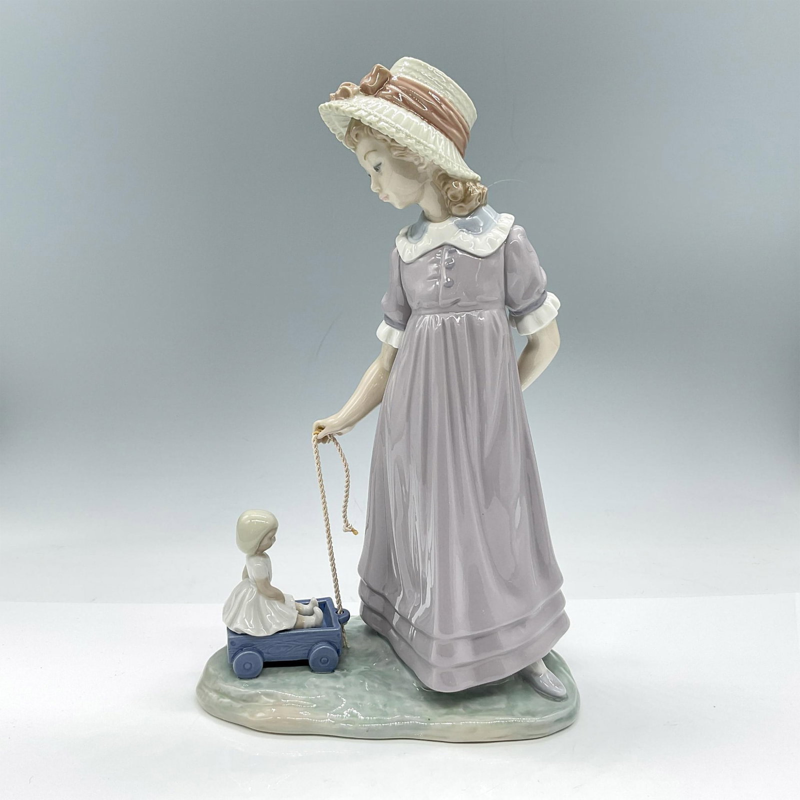 Girl with Toy Wagon 1005044 - Lladro Porcelain Figurine: Glossy handmade porcelain crafted in pale pastel colors. Depicts a childhood scene, a girl dressed in a lavender dress with a white collar and a wide-brimmed hat adorned with a soft pink ribbon, is at