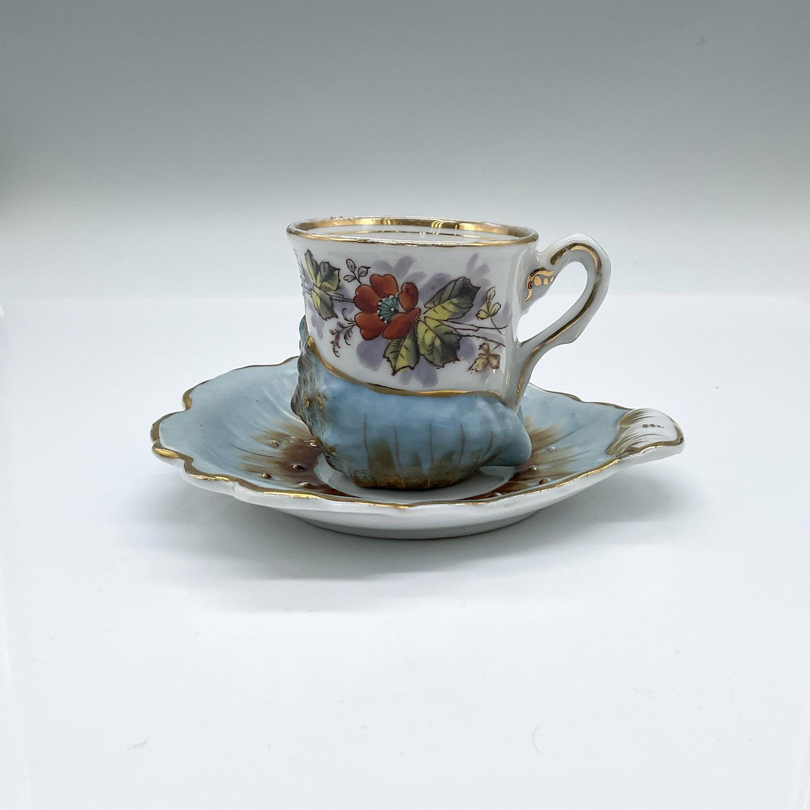 Unique Porcelain Demitasse Cup and Saucer: An unique but beautiful espresso set of the cup and saucer formed as a blue seashell with gilt accents. Hand painted flowers adorns the cup. Cup: 3.25"L x 2.5"W x 2.5"H. Saucer: 5.5"L x 5.25"W x 0.75"