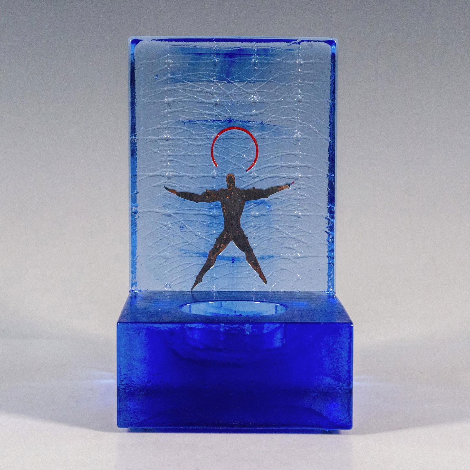 Kosta Boda by Bertil Vallien Blue Art Glass Candle Holder: A sculpted blue tint glass votive candle holder featuring an illustration of a man under the rising sun. Kosta Boda marking on the underside. Artist: Bertil Vallien Dimensions: 4"L x 3.25"W x 5.5"H Ma