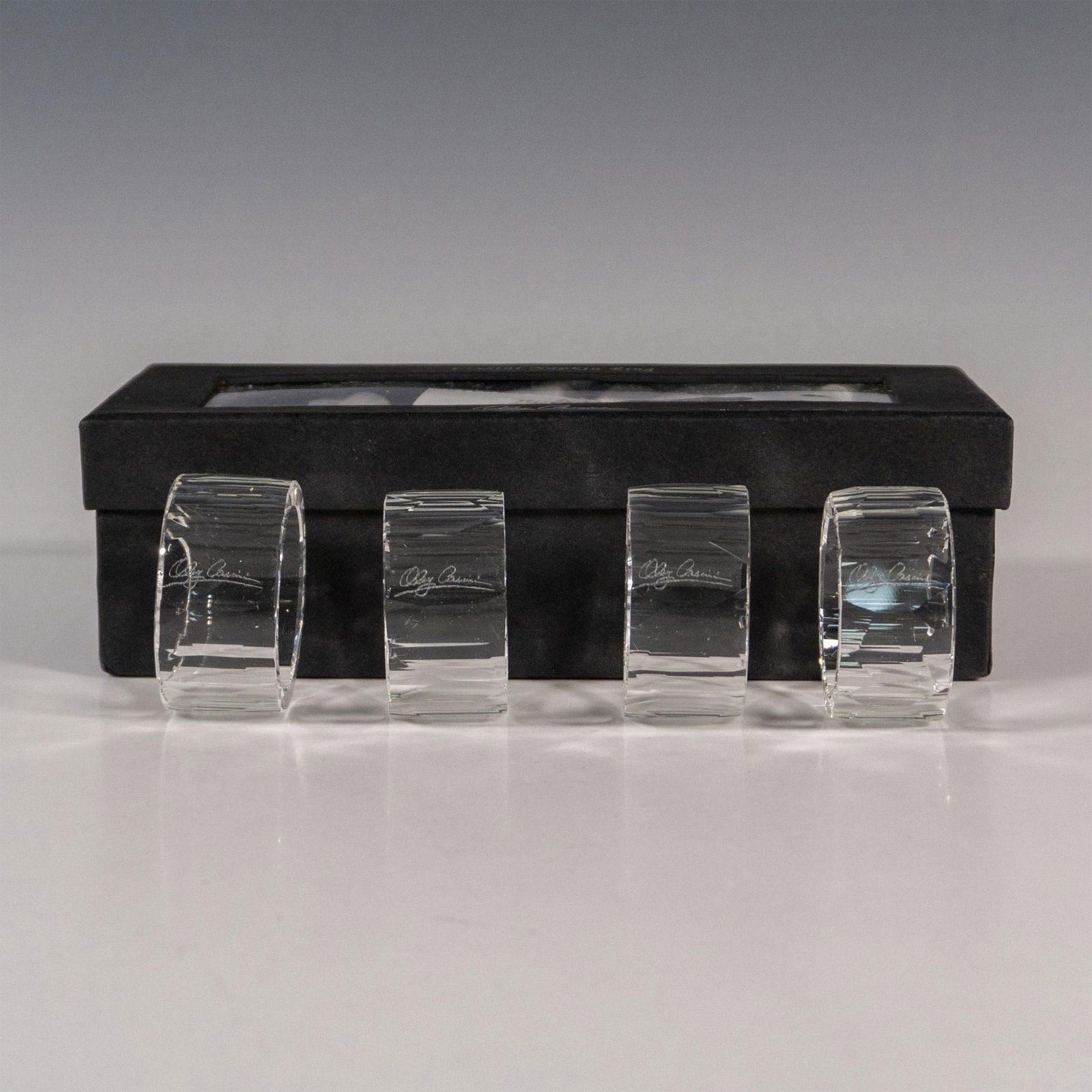Oleg Cassini Crystal Napkin Ring Holders, Jackie: A set of 4 clear round serviette rings with geometric cut panels. Oleg Cassini etched signature on each. This item has its original box: 8"L x 3.75"W x 2.5"H. Ring: 1"W; exterior: 2" dia; interior: 1.