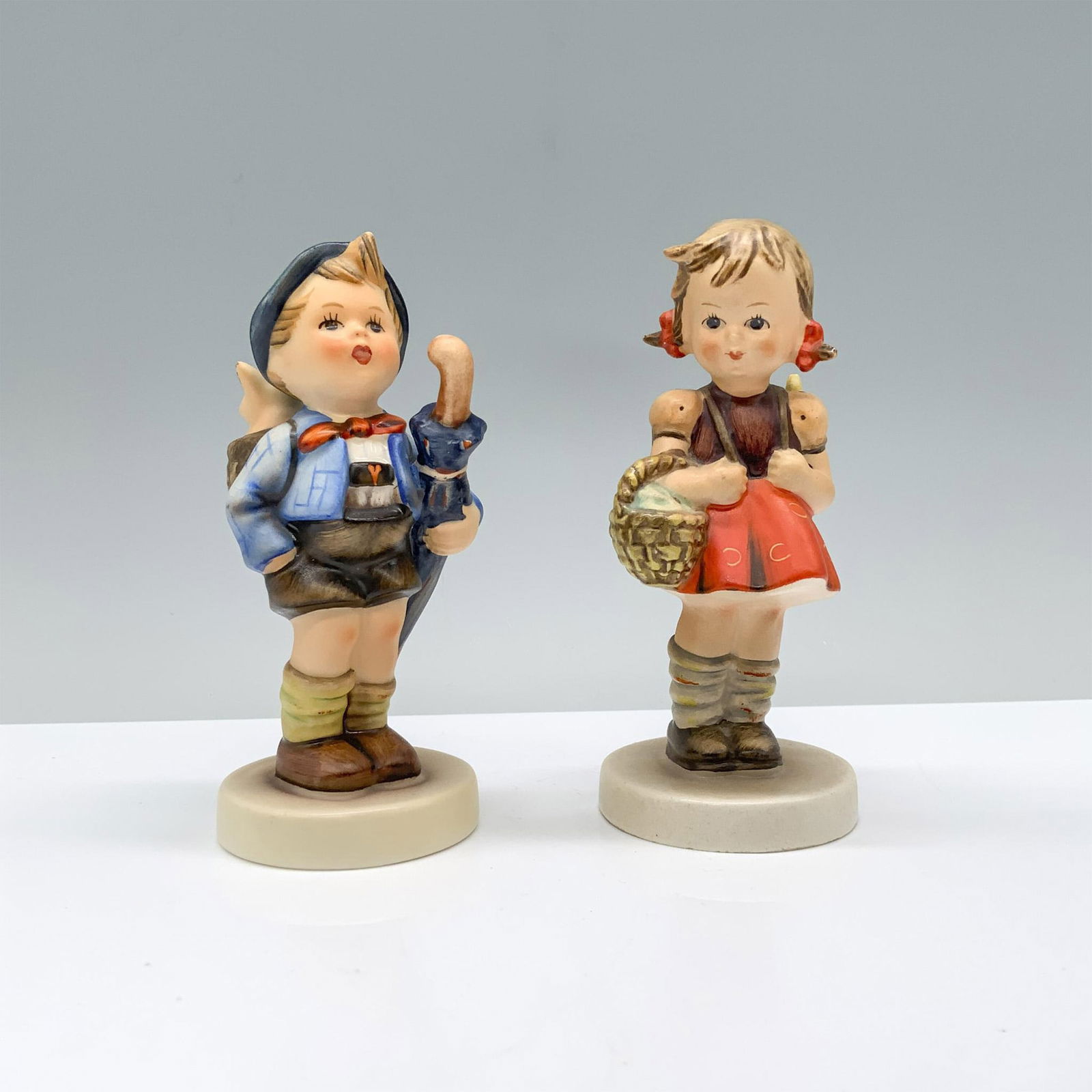 2pc Goebel Hummel Figurines, School Girl & Home From Market: A charming set featuring a young schoolgirl with a backpack and hand basket, accompanied by a young boy holding an umbrella with his pet pig in a basket on his back. Dimensions of the largest item: 2.