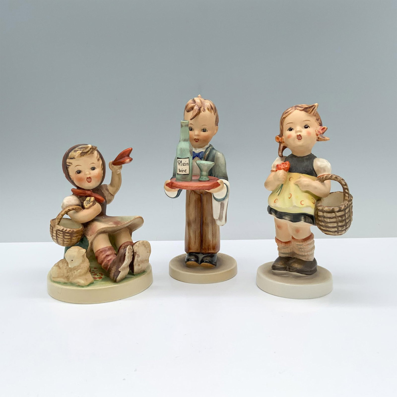 3pc Goebel Hummel Figurines, Sister Girl, Waiter, Farewell: A sweet girl sitting with a baby sheep, a young waiter boy serving a bottle of wine with flair, and an adorable girl standing with a flower and basket. Goebel Hummel backstamp. Dimensions of the large