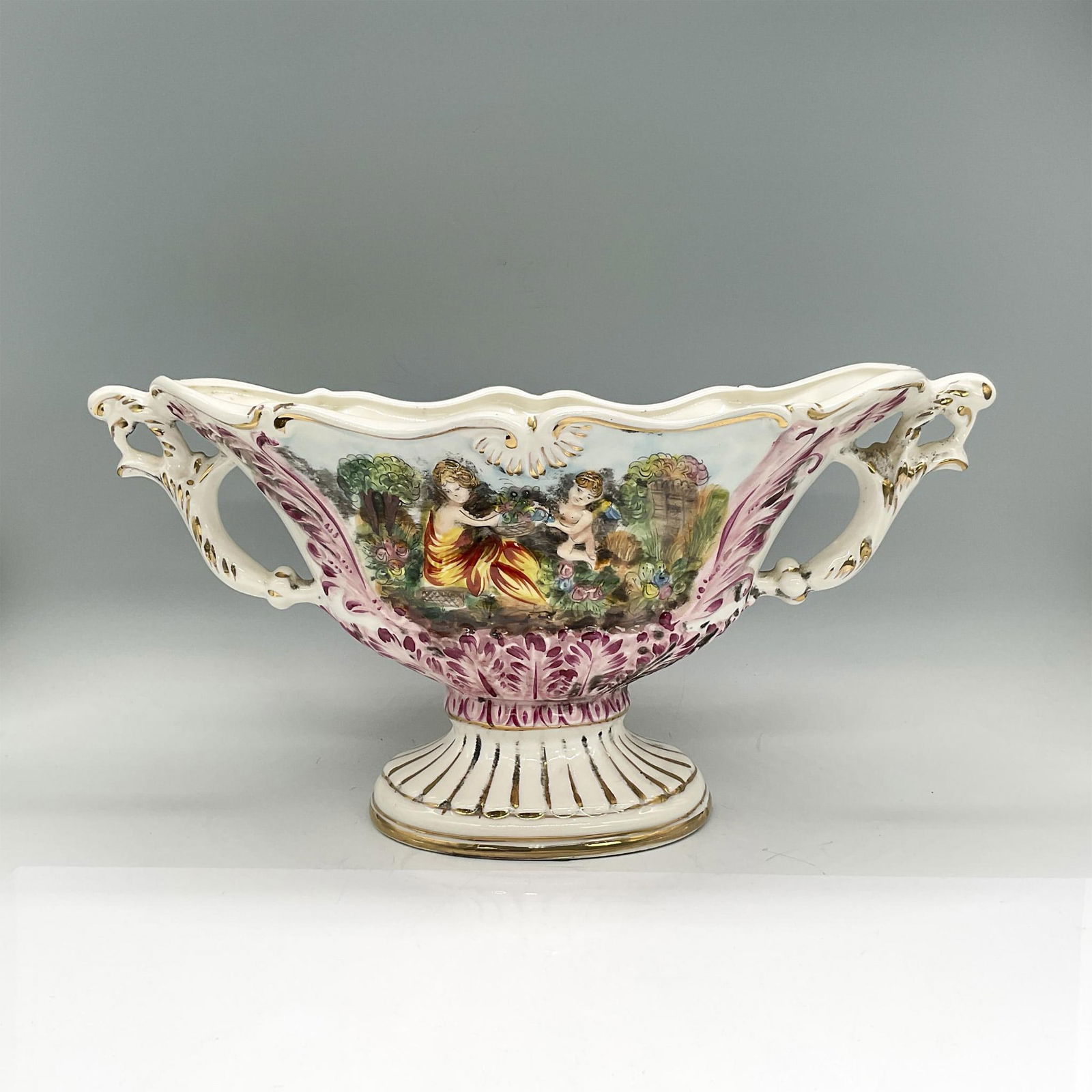 Capodimonte Pedestal Vase, In The Garden: Wide handpainted porcelain vase with two scenes: a lady with a cherub another of partially clothed ladies enjoying the outdoors; gold accents. Capodimonte mark. Dimensions: 15"L x 6.75"W x 7.25"H Manu
