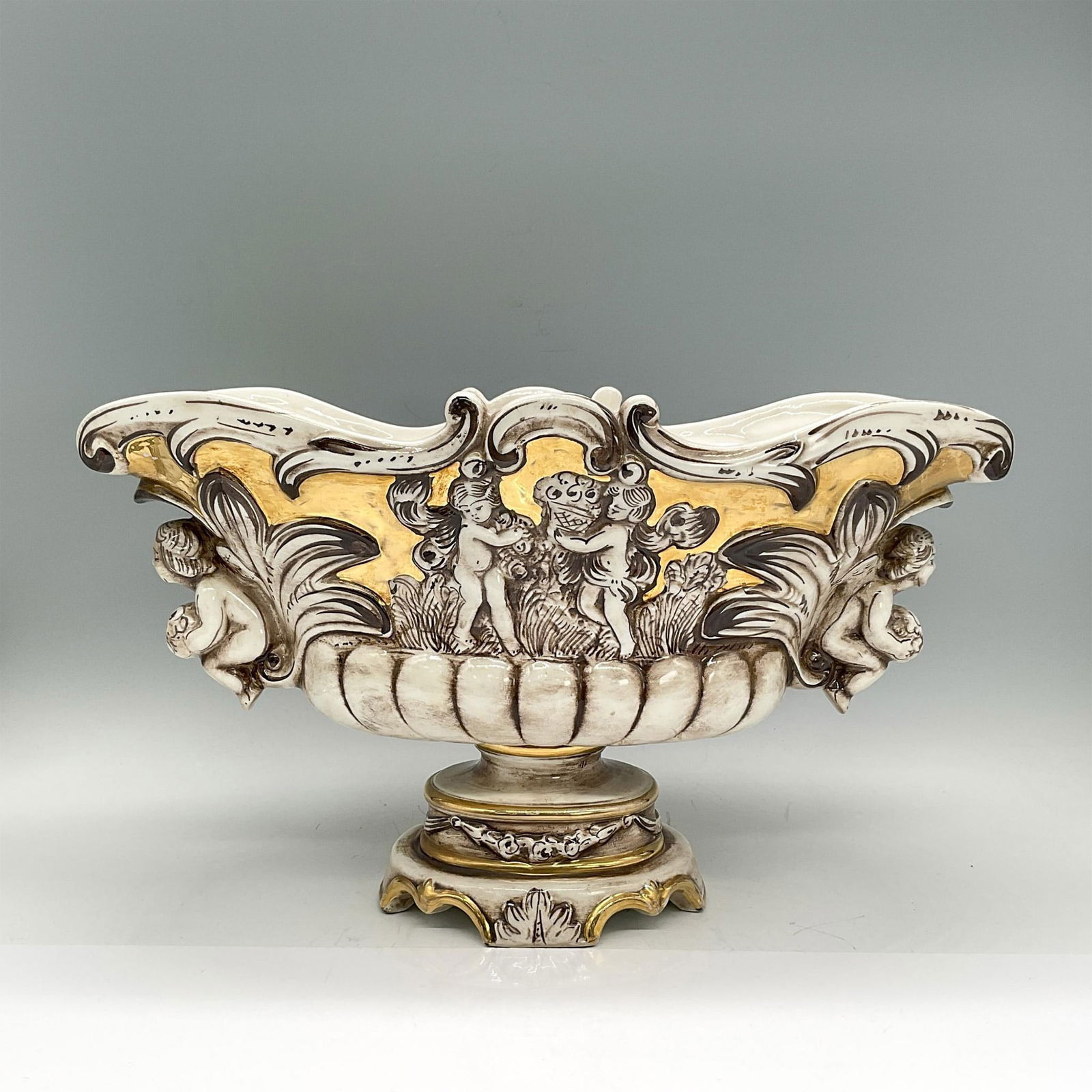 Capodimonte Pedestal Vase, Cherubs: Handpainted oval vase with a design of various nude people in high-relief, brown, white and gold coloration. Capodimonte mark. Dimensions: 14"L x 5.75"W x 8"H Manufacturer: Capodimonte Country of Orig