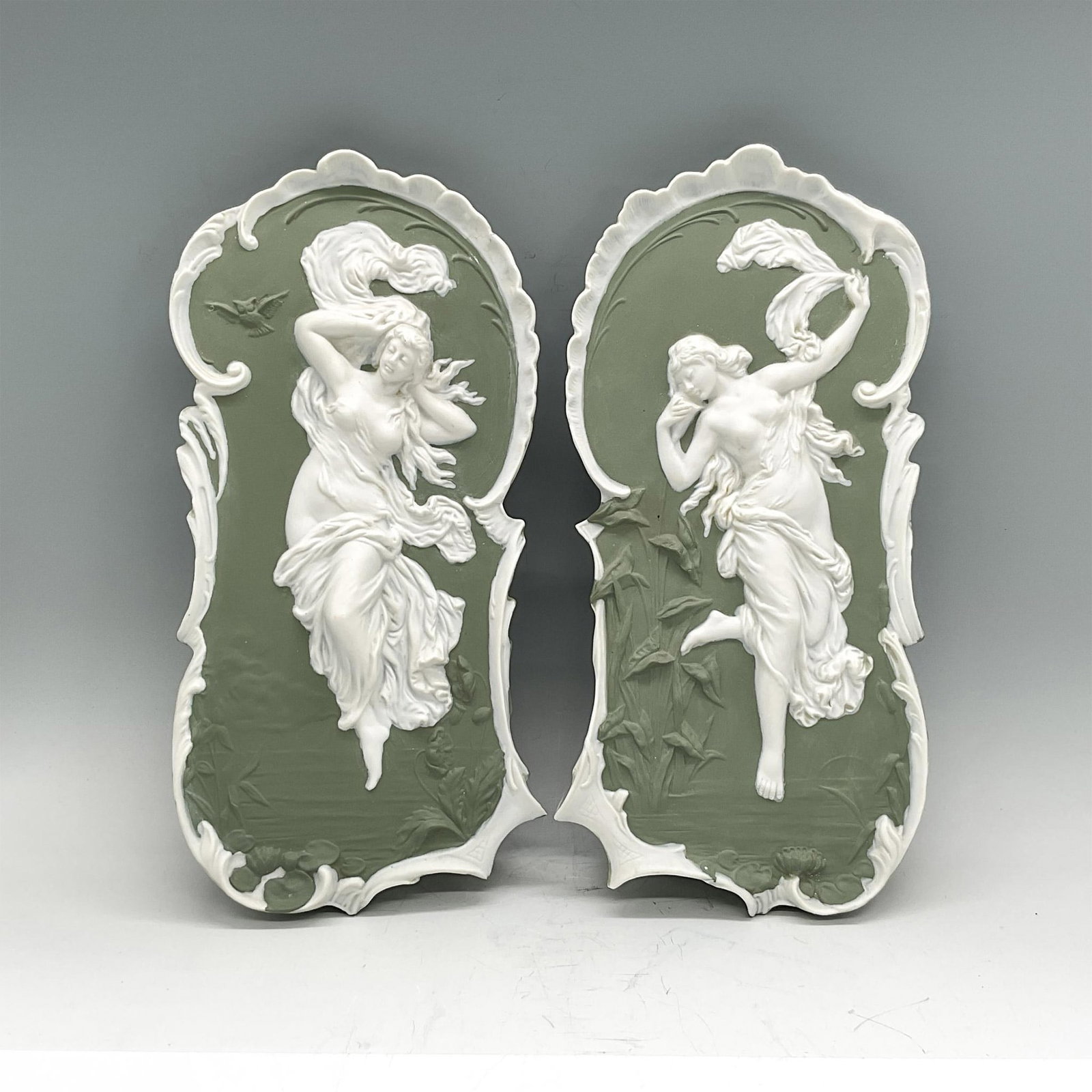 2pc Neoclassical Sage Green Wall Plaques, Nude Ladies: Two sage green stoneware wall plaques with sprigged images of nude ladies adorned with flowing fabrics in high-relief. In the style of Wedgwood Jasperware. Dimensions: 12.25"H x 5.25"W x 1.25"D Editio