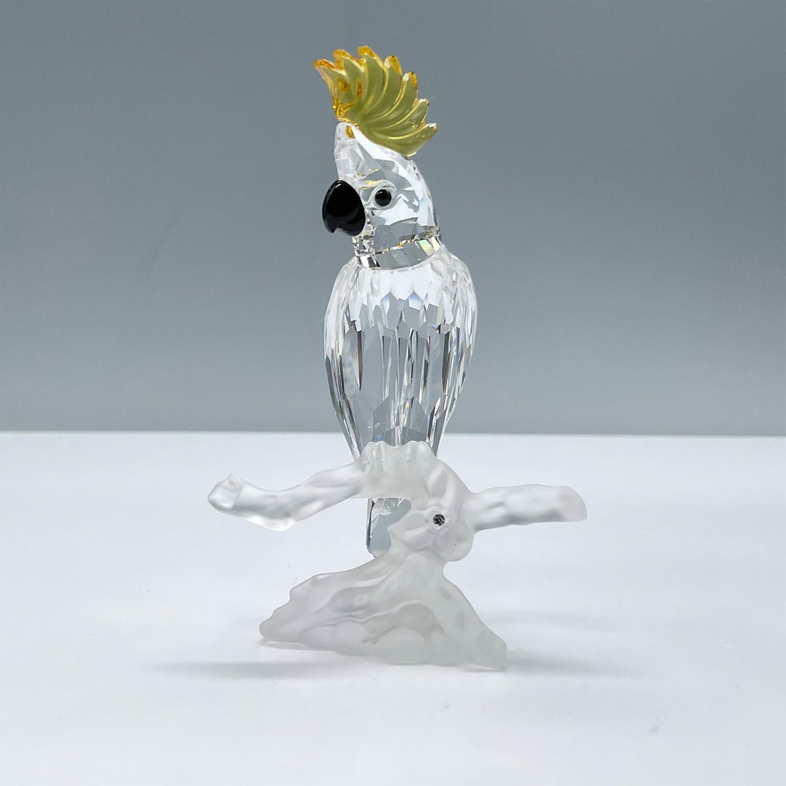 Swarovski Crystal Figurine, Cockatoo 261635: Beautiful clear crystal cockatoo bird with a yellow crest, black beak and eyes. It is sitting on a frosted crystal branch. Includes the original box, dimensions: 6.5''H x 3.25''dia. Swarovski etched b