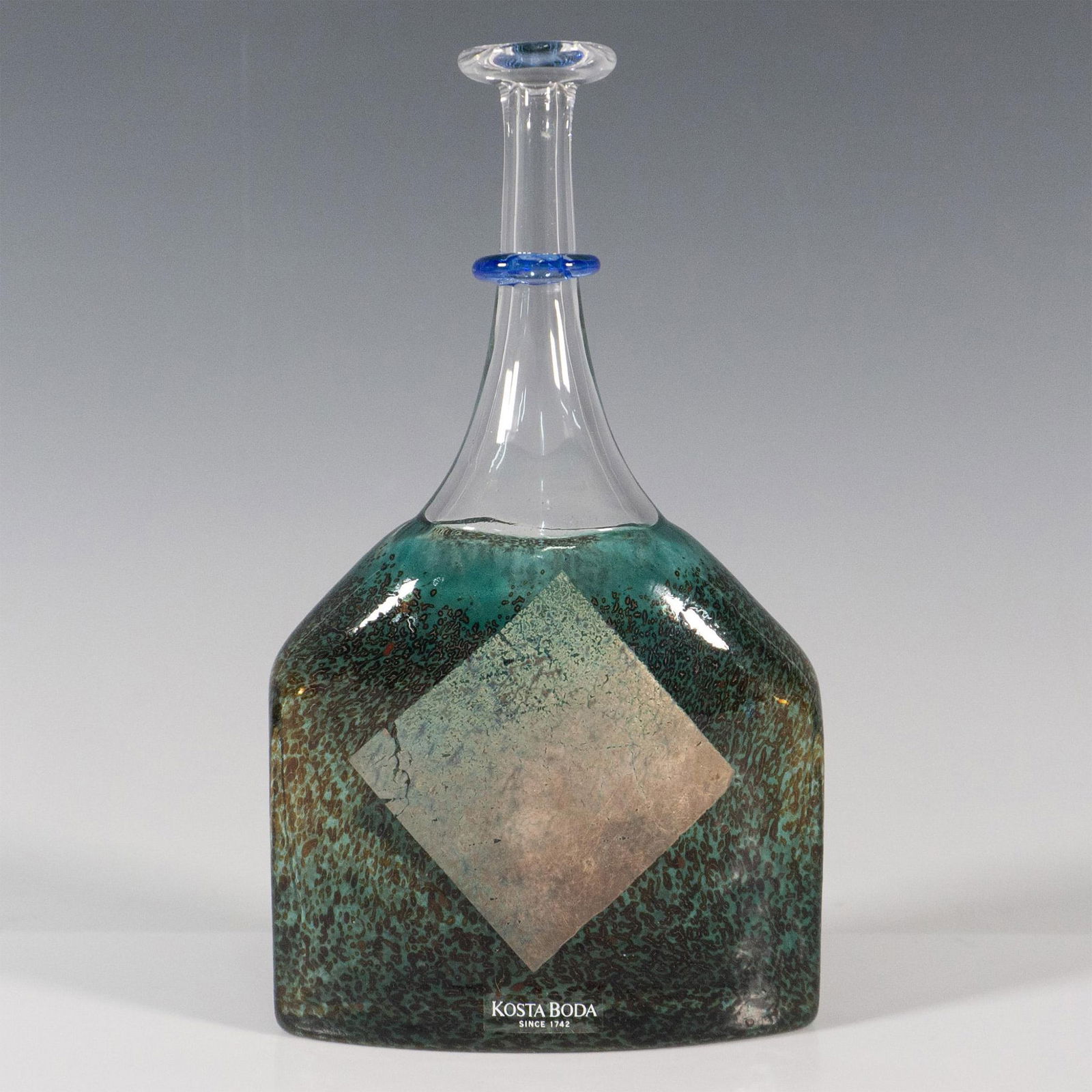 Kosta Boda Bottle Vase, Satellite: Beautiful crystal accented by a blue glass ring and decorated in a rich green textured body. Kosta Boda engraved signature. Artist: Bertil Vallien Dimensions: 1''L x 4''W x 7''H Manufacturer: Kosta Bo