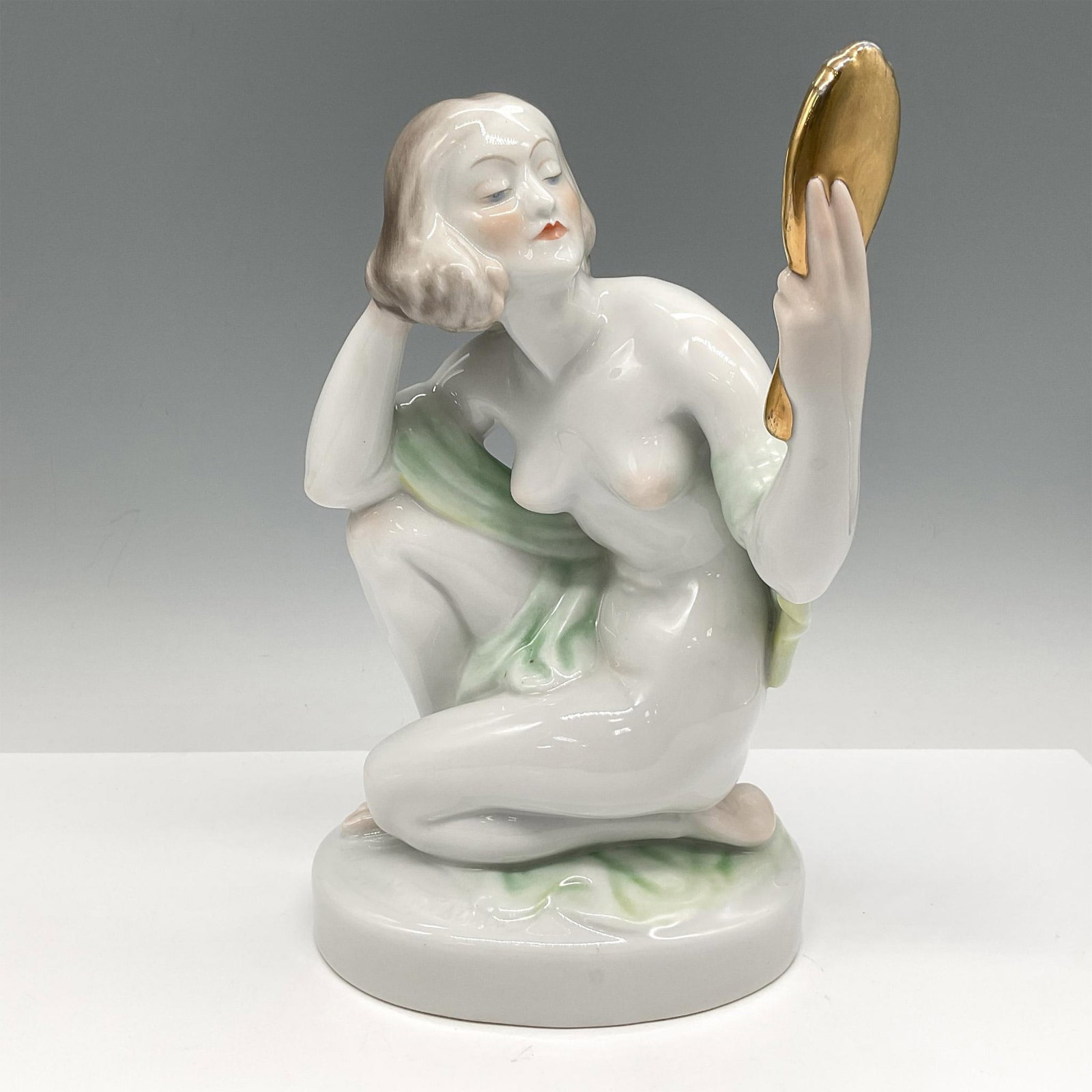 Herend Porcelain Figurine, Art Deco Style Nude: Glossy figural admiring her image in the mirror. Sitting nude with green sheath around her shoulders. Herend backstamp. Issued: 20th century Dimensions: 5.5"L x 6"W x 9.25"H Manufacturer: Herend Count
