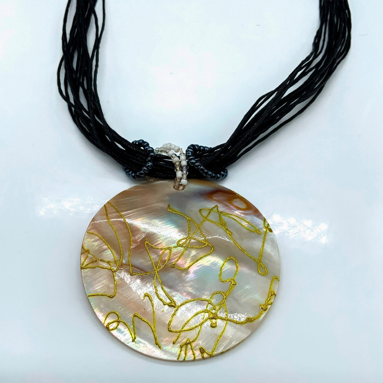 Hand-Painted Mother Of Pearl Medallion Pendant Necklace: Large round mother of pearl medallion pendant decorated with golden swirls measuring 2.5"dia. The necklace is multiple strands of black cord artfully wrapped at the top with a button clasp. Dimensions