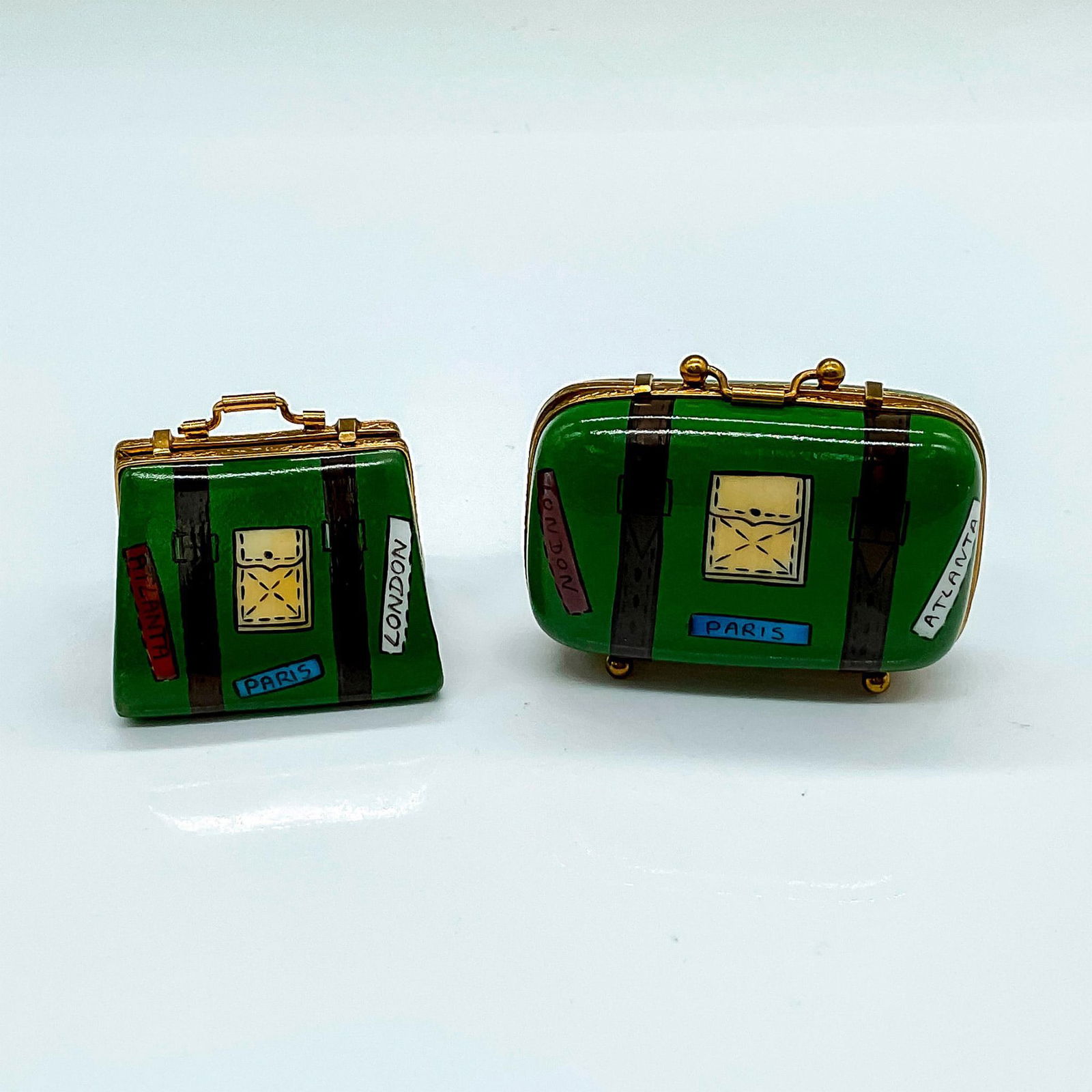 Pair of Limoges SE Porcelain Suitcase Boxes: A set of two mini hand painted green suitcases with travel stickers on the front. Pink and green spotted pattern on the interior. Limoges France Peint Main SE on the inside. The largest piece measures