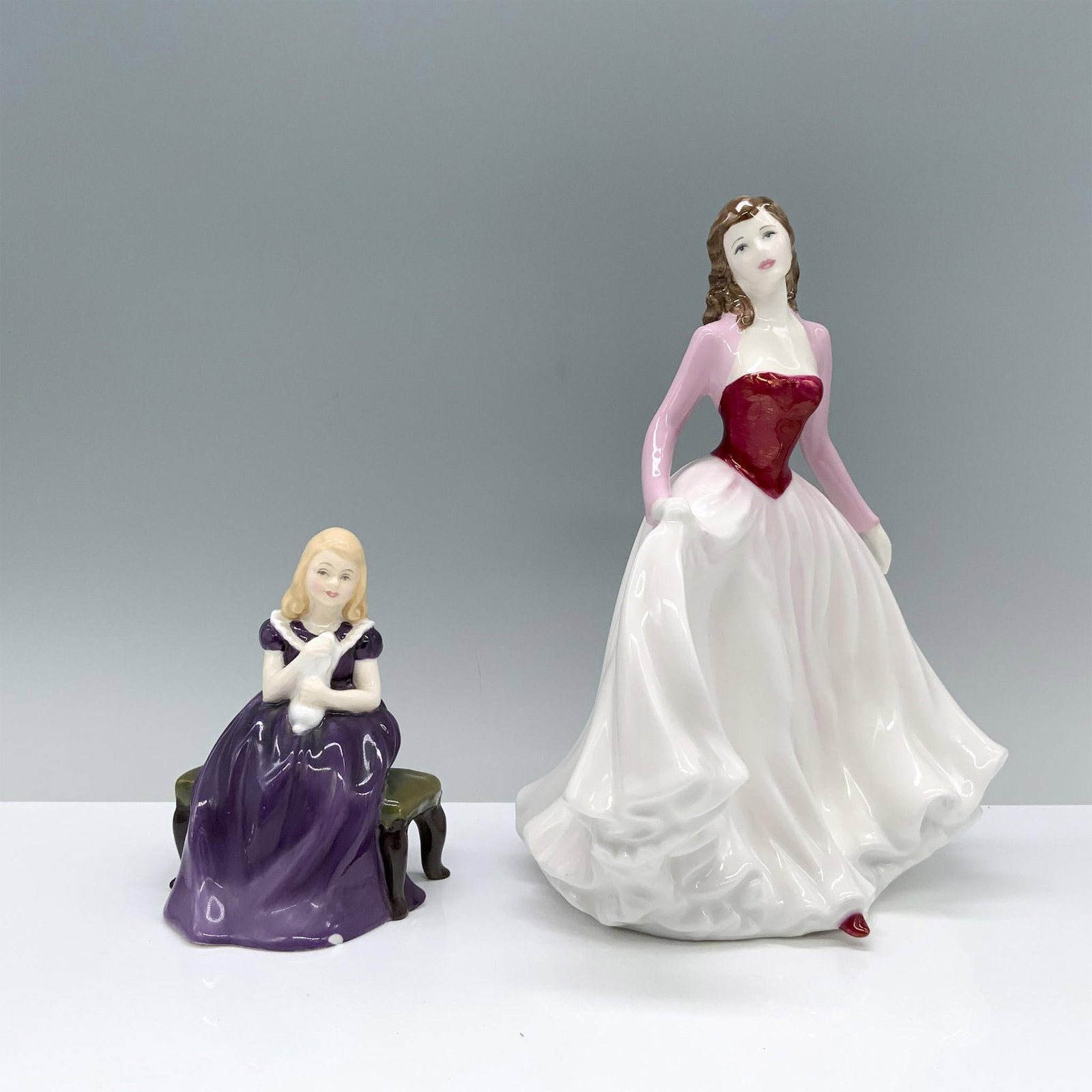 2pc Royal Doulton Figurines, Affection HN2236, Pink HN4606: A young blonde girl in a purple dress tenderly cradles her pet close and a brunette woman in pink, red, and white gracefully performs a curtsey. Royal Doulton backstamp. Dimensions: 3.25"L x 2.75"W x 