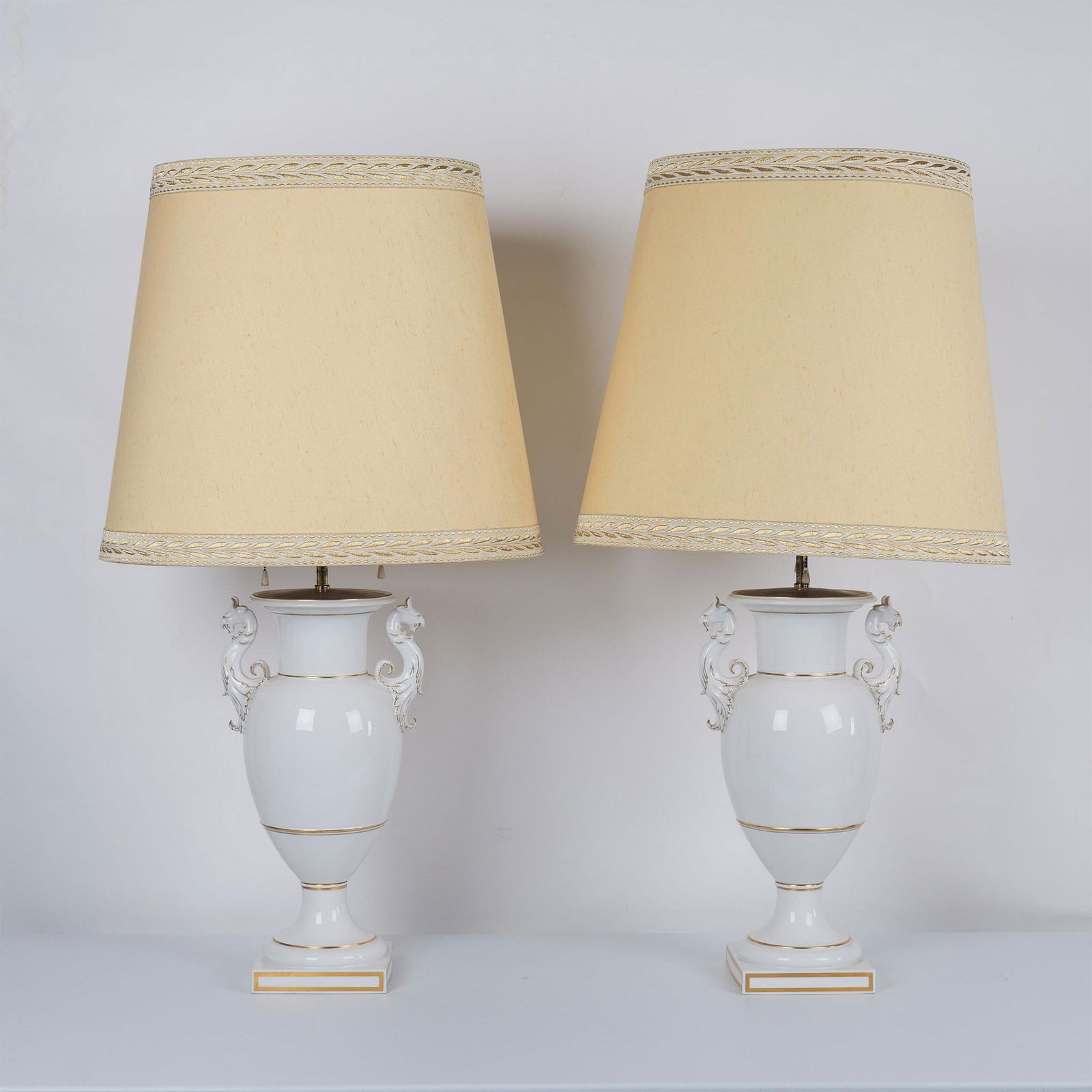 Pair of KPM White Porcelain Amphora Lamps: Adorned with griffons on each side with gilt accents throughout. Brass mounts hold pull chain lights. Large fabric shades with embroidered banding is included. Issued: 20th century Dimensions: Lamp 10