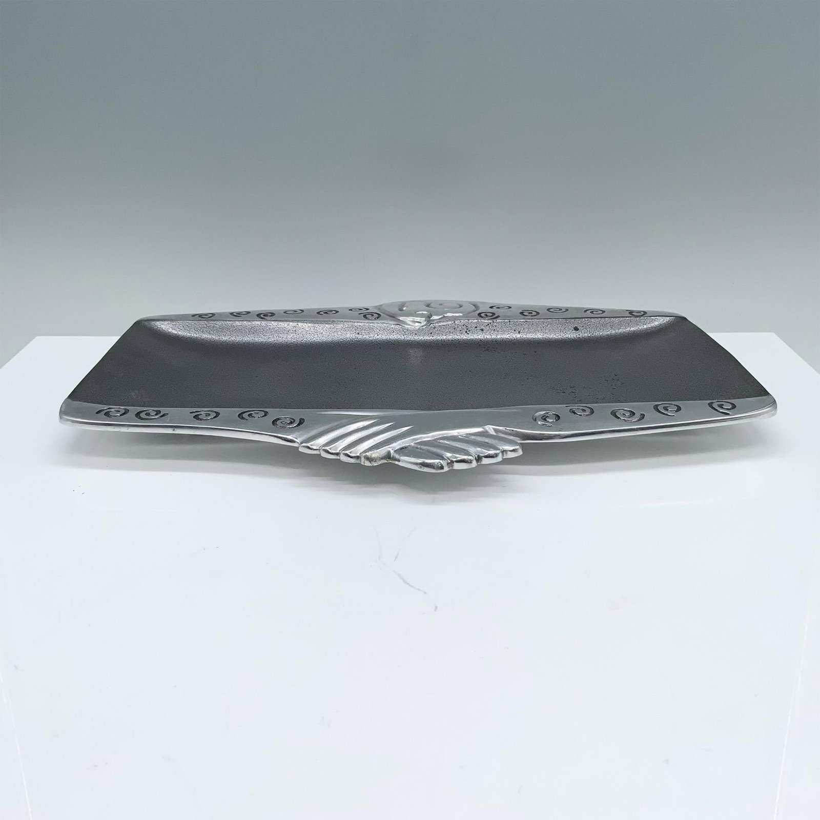 Carrol Boyes Serving Platter, Kimono: A silver-colored platter adorned with swirling patterns and featuring two interlocking hands at the bottom of its rim. Marked Carrol Boyes. Artist: Carrol Boyes Dimensions: 12.75"L x 9.5"W x 0.5"H Man