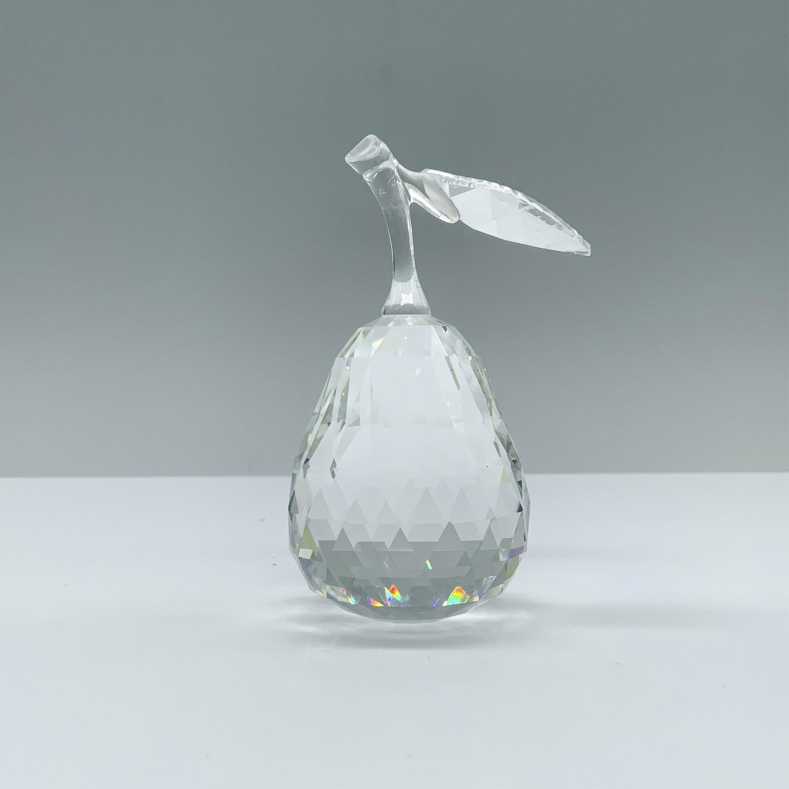 Swarovski Crystal Figurine, Pear 162885: A stunning clear pear featuring triangular cuts and a smooth stem, elegantly crowned with a leaf at the top. Swarovski backstamp. This item has its original box. Dimensions of the box: 4.75"H x 6.25" 