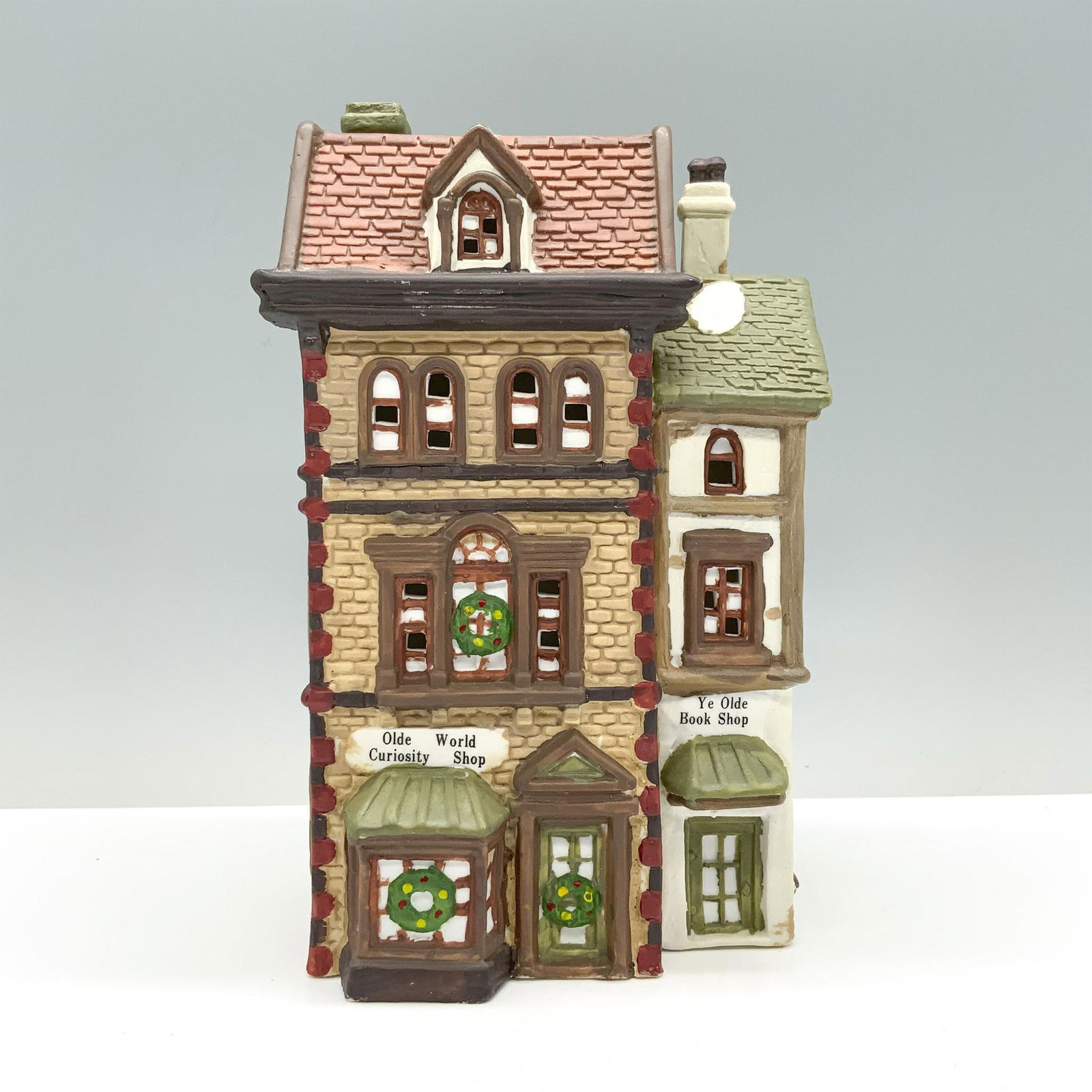 Dickens Keepsakes House Figurine, Olde World Curiosity Shop: A shop adorned with beige brick walls, a red tile roof, and open windows accented with wreaths, that also features a quaint white bookshop attached to its side. Dimensions: 4.5"L x 3.75"W x 8.5"H Manu
