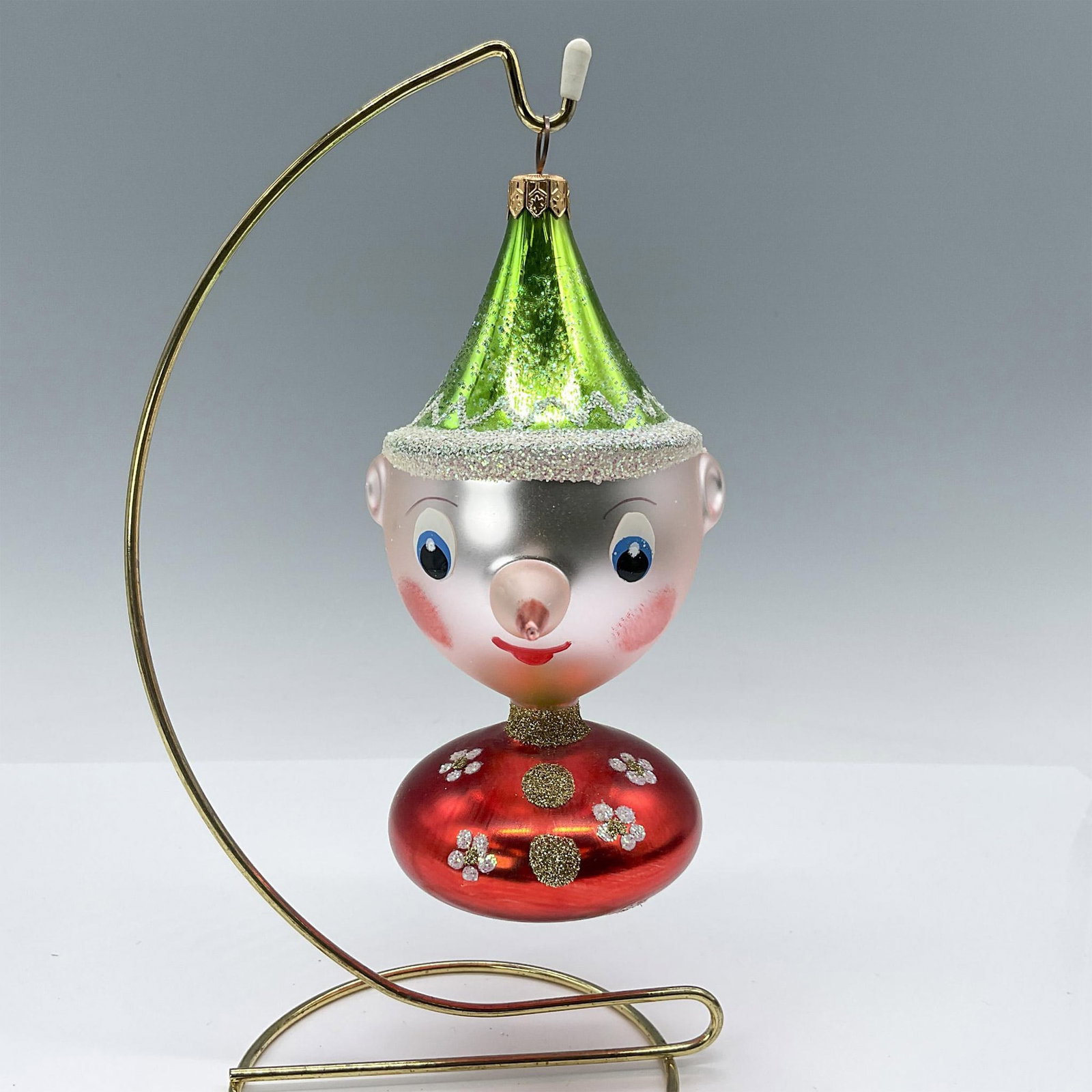 Christopher Radko Italian Blown Glass Ornament, Pinocchio: Beautiful hand-painted and mouth-blown ornament depicting Pinocchio with his long pink nose, dressed in a red top and green hat adorned with glitter. Issued: 1993 Dimensions: 3"L x 4.5"W x 6.75"H Manu