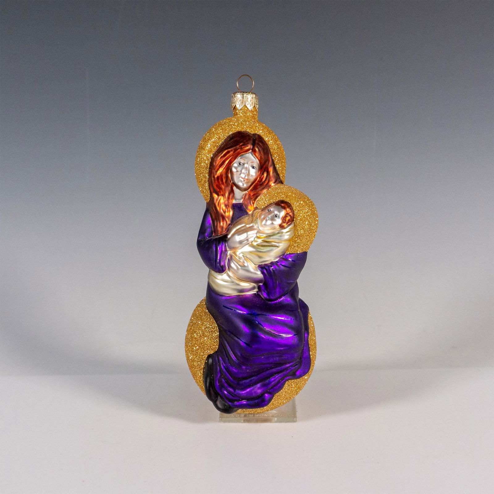 Patricia Breen Christmas Ornament, Seated Madonna: This exquisite glass ornament boasts a European-style hand-painted design portraying Madonna with child, adorned in rich gold and majestic purple hues, accented with shimmering glitter to captivate th
