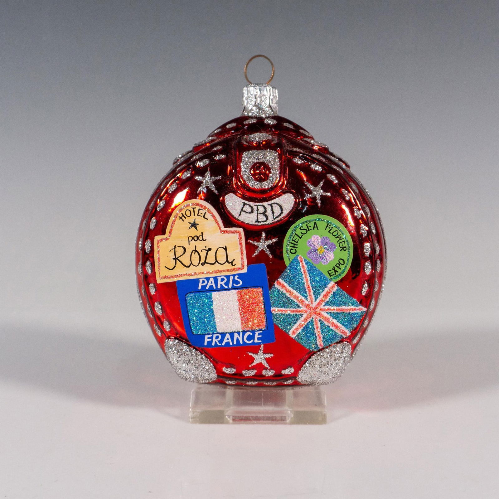 Patricia Breen Christmas Ornament, Journey With Me: Exclusively available at Lowe Art Museum in Coral Gables, this exquisitely hand-painted European-style glass ornament depicts a red suitcase adorned with colorful stickers on the exterior, including o
