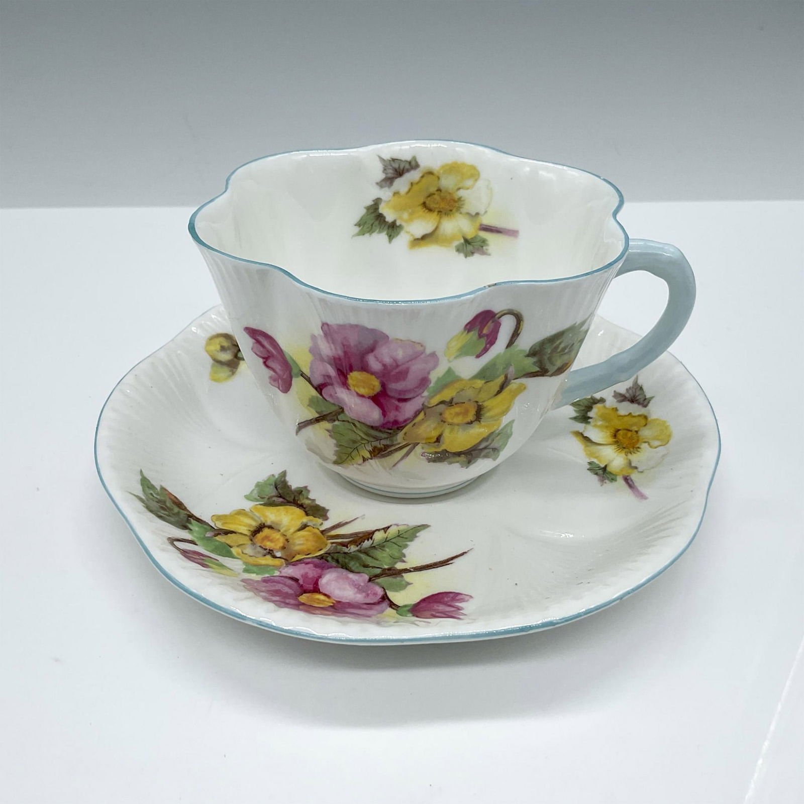 Shelley Bone China Tea Cup and Saucer, Begonia: This beautiful fine Bone China set is embellished with hand painted colorful flowers and blue accents. Shelley backstamp. Issued: 20th century Dimensions: Cup: 3.5''L x 4''W x 2.5''H; Saucer: 0.75''H