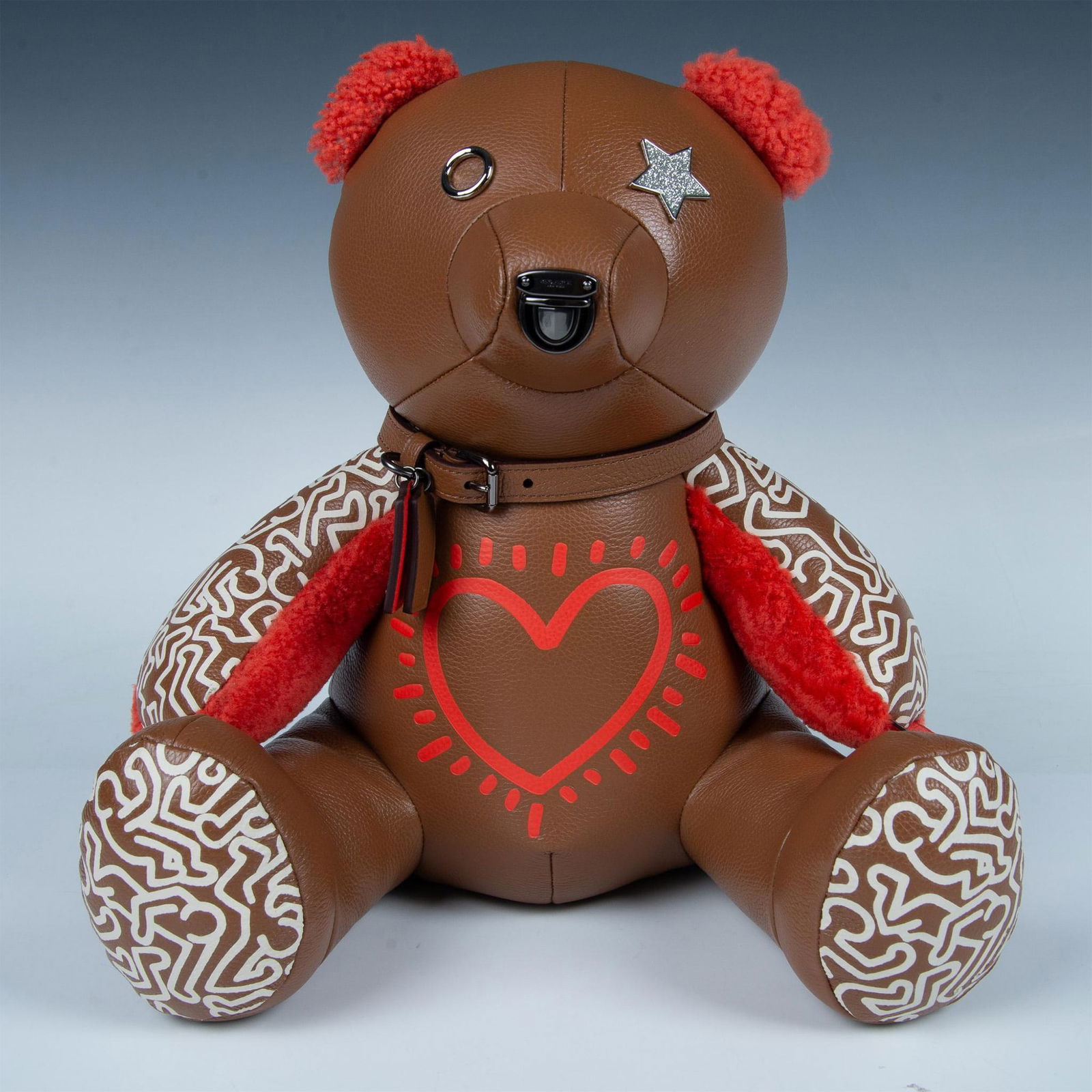 Coach Keith Haring Collaboration Plush Leather Teddy Bear: Brown leather teddy bear with orange plush details and iconic Keith Haring chalk outlines on the outside of paws and bottom of feet. Classic heart outline in center of chest. Coach buckle nose, silver