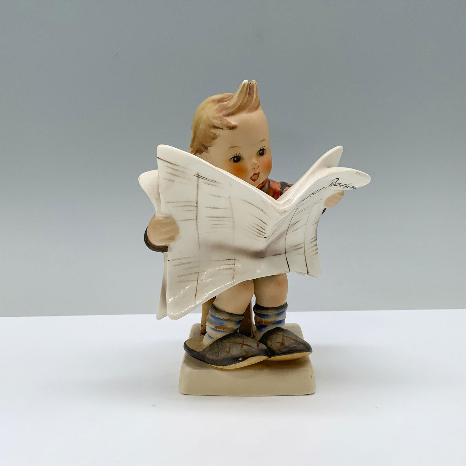 Goebel Hummel Porcelain Figurine, Latest News: Perched on a stool, a young boy, snug in an orange jacket, slips his feet into his parents oversized slippers, absorbed in reading his daily news. Matte finish. Goebel backstamp. Dimensions: 4"L x 2.5