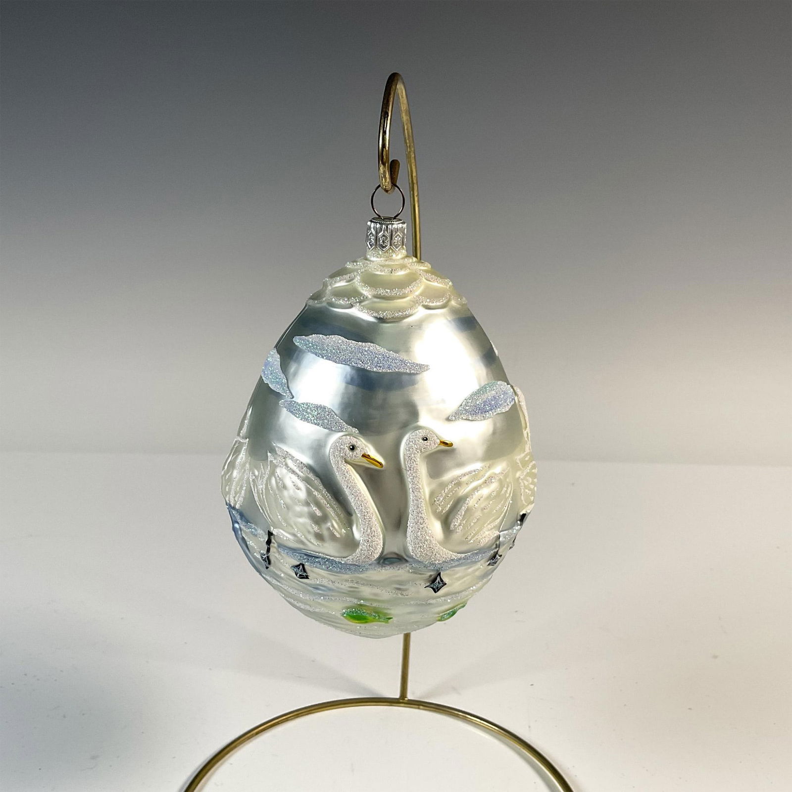 Patricia Breen Christmas Ornament, Seven Swimming Swans: This mouth-blown and hand-painted glass ornament takes the shape of a large egg and depicts seven graceful swans swimming in a serene lake. Adorned with blue hues and embellished with glitter. Issued: