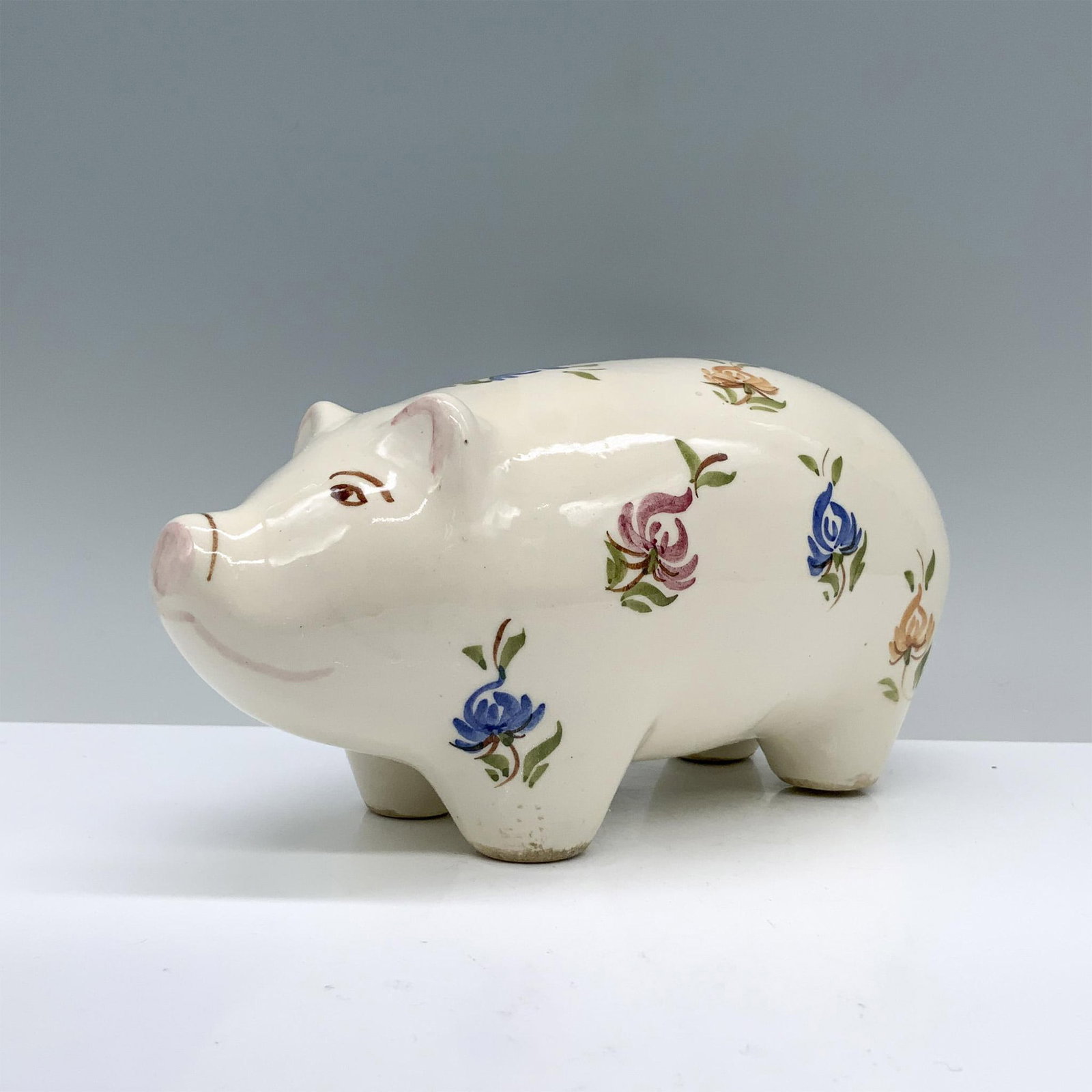 Cash Family Art Pottery Piggy Bank: A cream-colored piggy bank adorned with charming blue, yellow, and purple flowers delicately painted across its body. Marked Cash Family. Dimensions: 6.75"L x 3.25"W x 3.5"H Manufacturer: Cash Family 