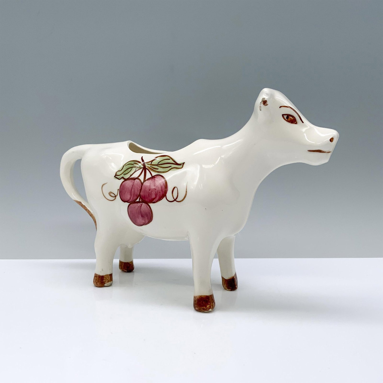 Clinchfield Art Pottery Creamer, Cow: A whimsical white cow-shaped creamer with elegant brown accents and charming cherries hand-painted on her body. Clinchfield backstamp. Dimensions: 8.25"L x 2.5"W x 5.5"H Manufacturer: Clinchfield Coun