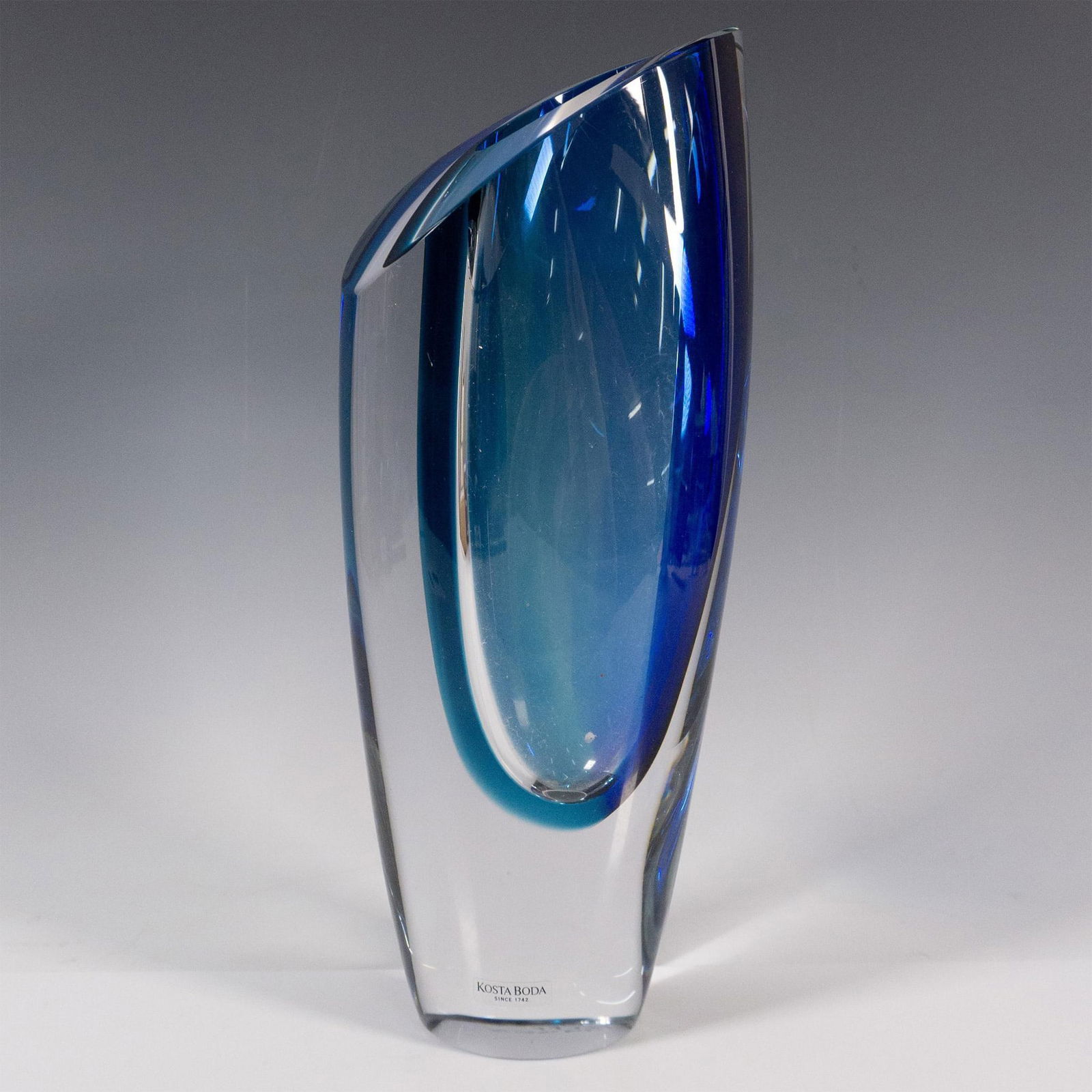 Kosta Boda by Goran Warff Blue Vase, Seaside: Clear with an angled surface, and decorated with a distinctive curved light blue and dark blue interior. Kosta Boda stickered. Artist signed on the underside. Artist: Goran Warff Dimensions: 4.5"L x 3