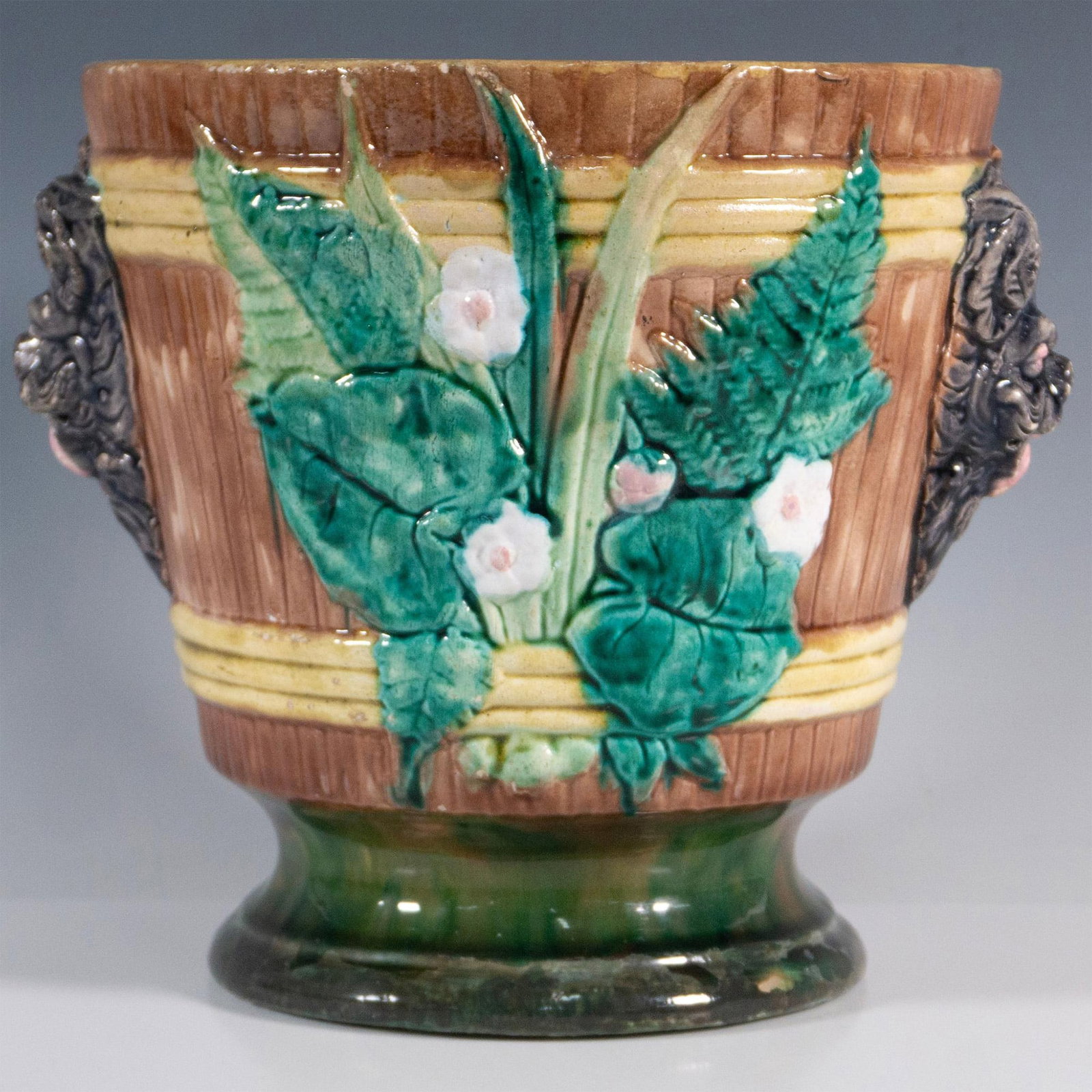 Majolica Jardiniere Mythical Figural Handles and Florals: A large bucket-shaped planter adorned with figural handles on either side and bas-relief botanical features between them. Hand-glazed in naturalistic hues of brown, greens, gold, and white, with a lig