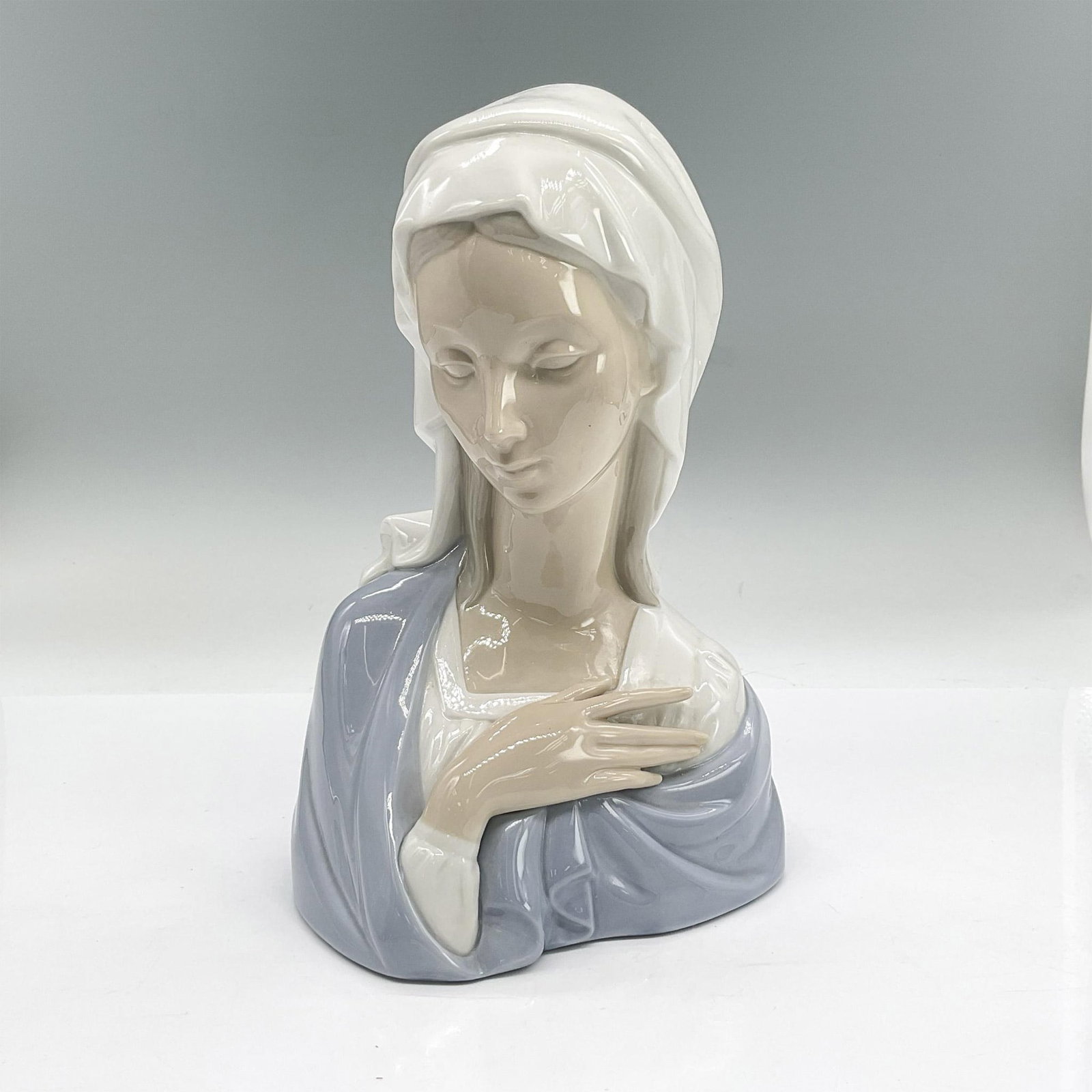 Lladro Porcelain Bust, Madonna Head 1004649: Glazed porcelain bust of the iconic Mary in a white headdress and blue shawl. Lladro backstamp. Issued: 1969-2001 Dimensions: 5.5"L x 3.75"W x 8.25"H Manufacturer: Lladro Country of Origin: Spain