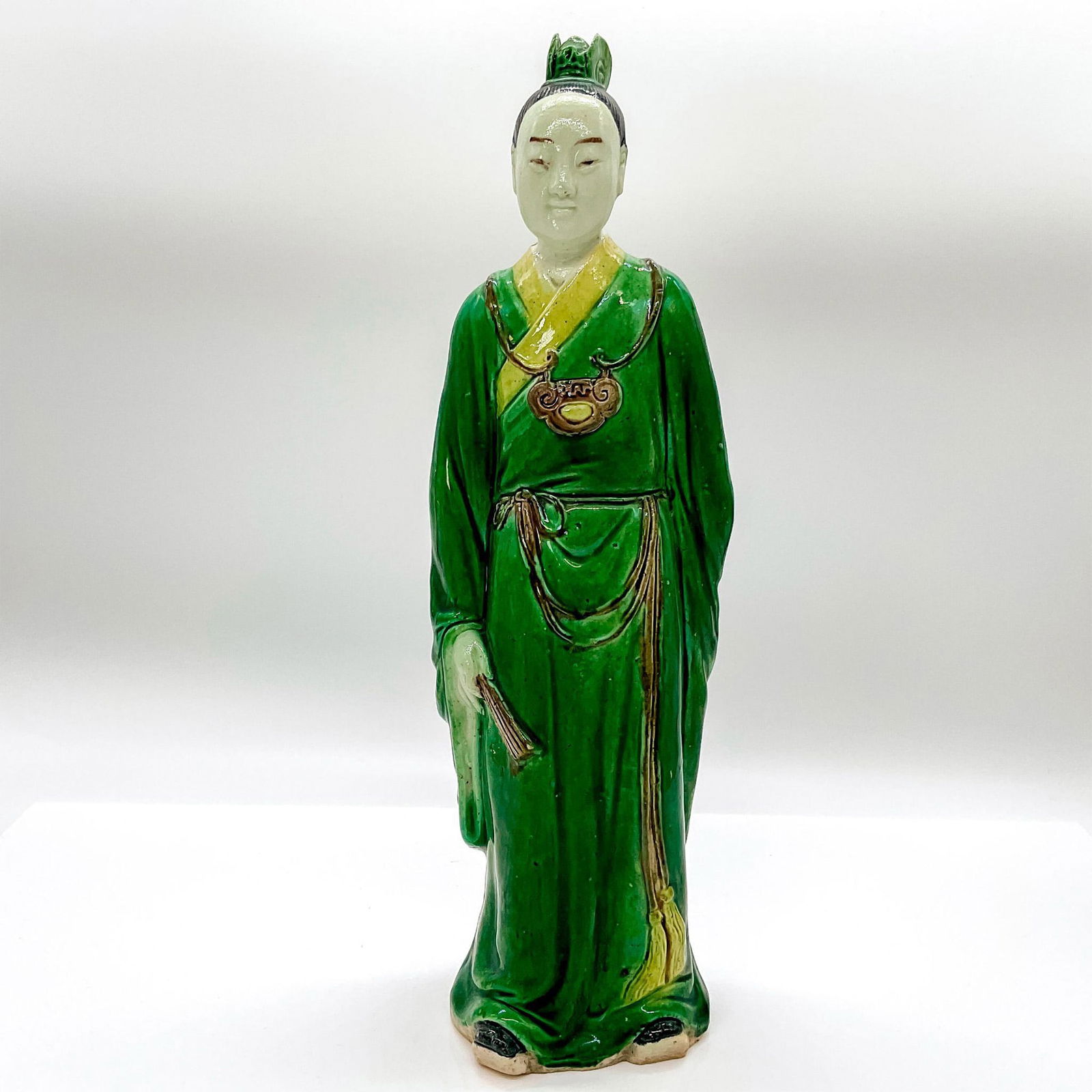 Chinese Ceramic Figure, Scholar: Hand painted tall figure of a scholar in a green robe with matching hairpiece, holding a hand fan. China and Chinese character marks impressed to bottom. Dimensions: 3.25"W x 11.5"H Country of Origin: