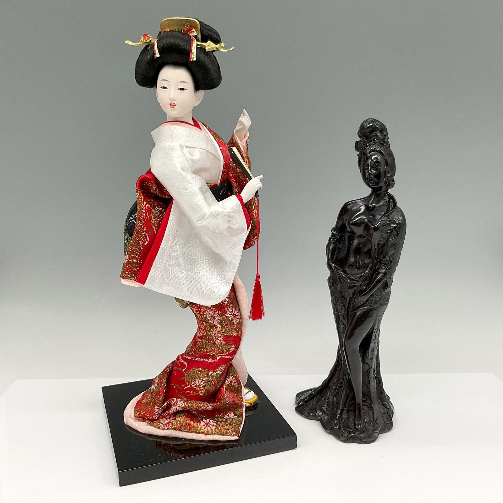 2pc Japanese Geisha Doll and Chinese Nude Figurines: Intricate replication of Geisha with colorful kimono, hair in traditional style, holding a fan with delicate detail to face and hands set on wood base 5"L x 5"W x 12.5"H. Carved cherry amber resin scu