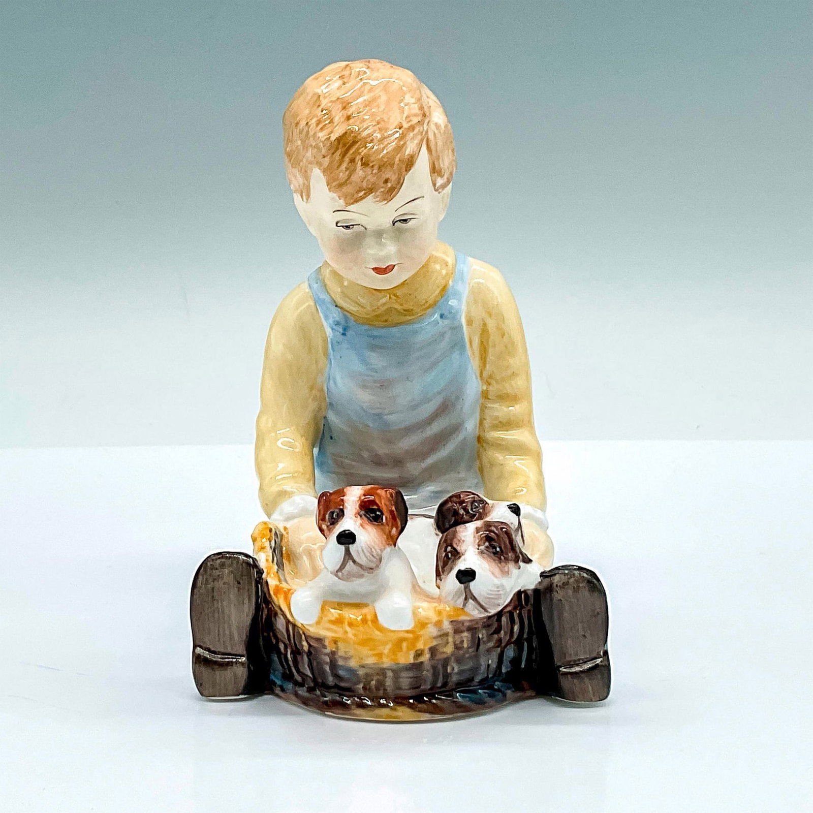Paragon Bone China Figurine, Pals: Painted figure of a young boy with a basket of puppies. Yellow and blue coloration. Paragon backstamp. Issued: 20th century Dimensions: 3.25"L x 3"W x 4"H Manufacturer: Paragon Country of Origin: Engl