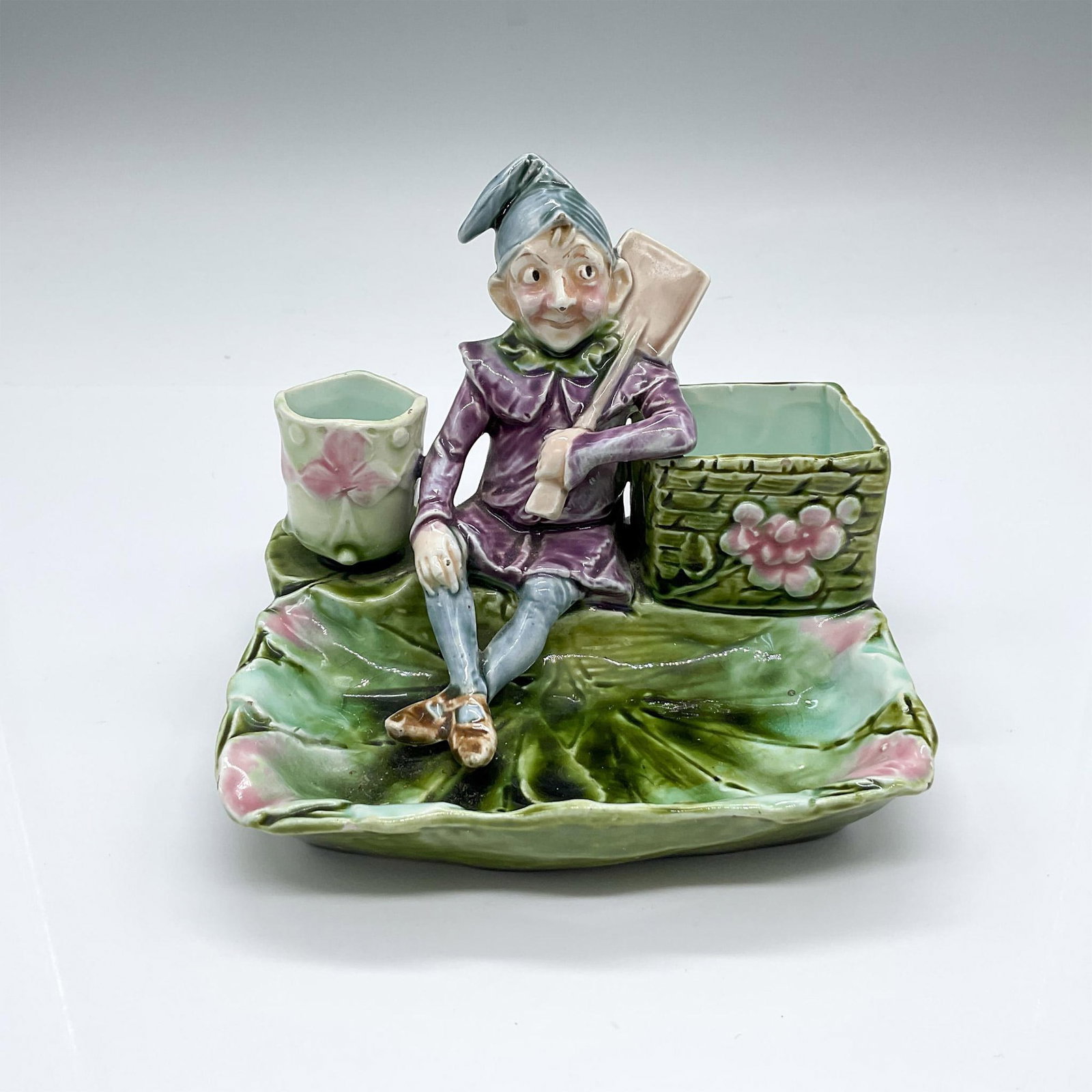 Majolica Pottery Elf Cigarette Holder: Hand made and hand painted tobacciana dish with a seated elf in purple holding a paddle, surrounded by pink flowers. Green, pink, purple, and blue colorations. Matchstick striker on the side. Dimensio