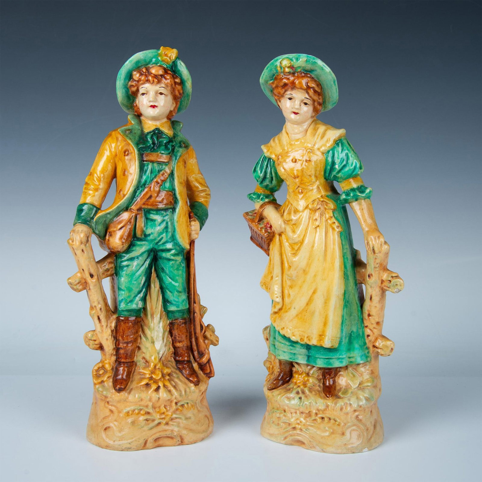 Pair of Majolica Pottery Girl and Boy Figurines: 2 hand made and hand painted figures of a young woman and man standing by a fence. The girl is holding a basket of flowers, and the man has a hunting rifle. Yellow, green, and brown colorations. Girl: