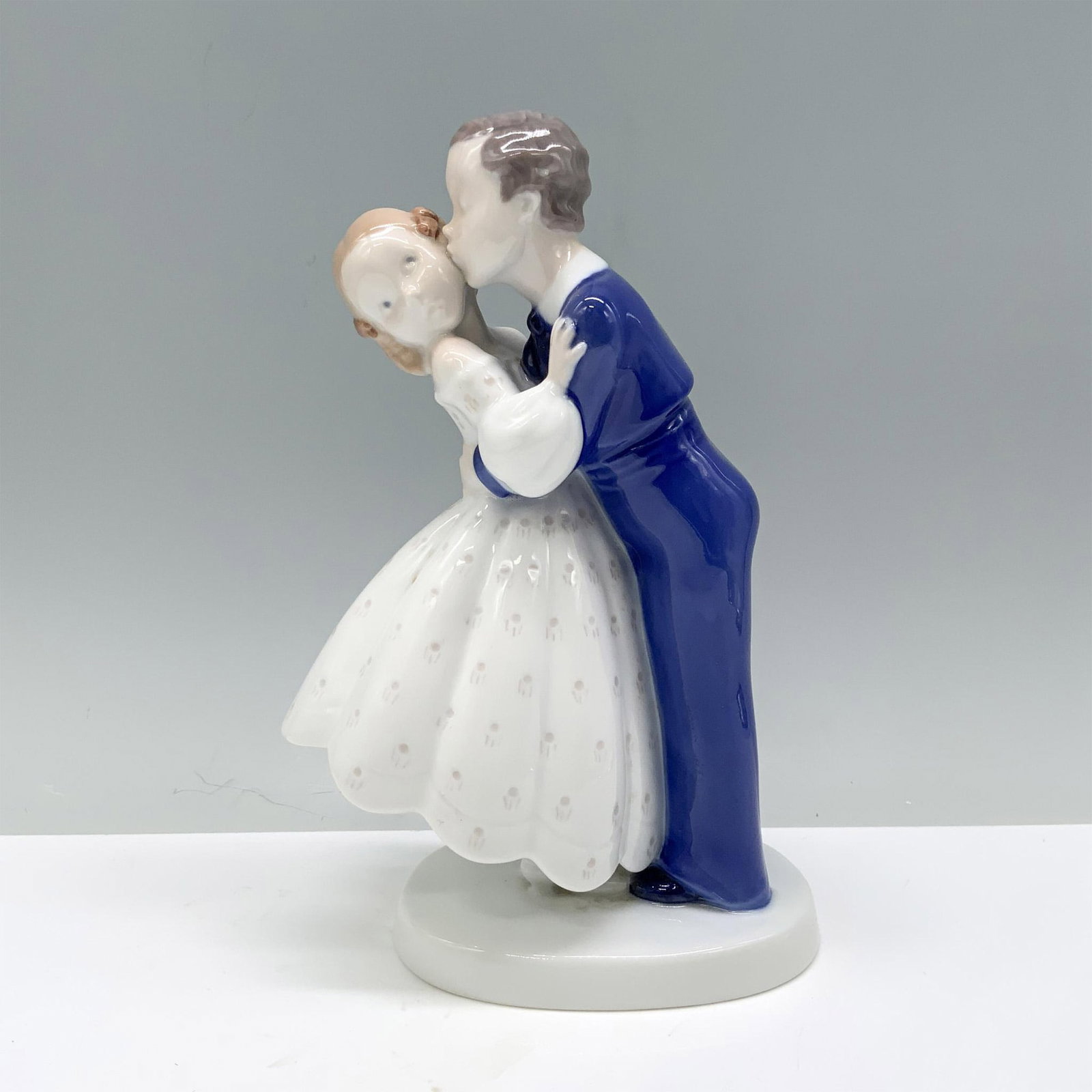 Bing & Grondahl Porcelain Figurine, First Kiss 2162: A young brunette man in a blue tux gently kisses his bride, who is in a white, flowing polka-dotted gown. B&G backstamp. Dimensions: 5.25"L x 3"W x 7.5"H Manufacturer: Bing & Grondahl Country of Origi