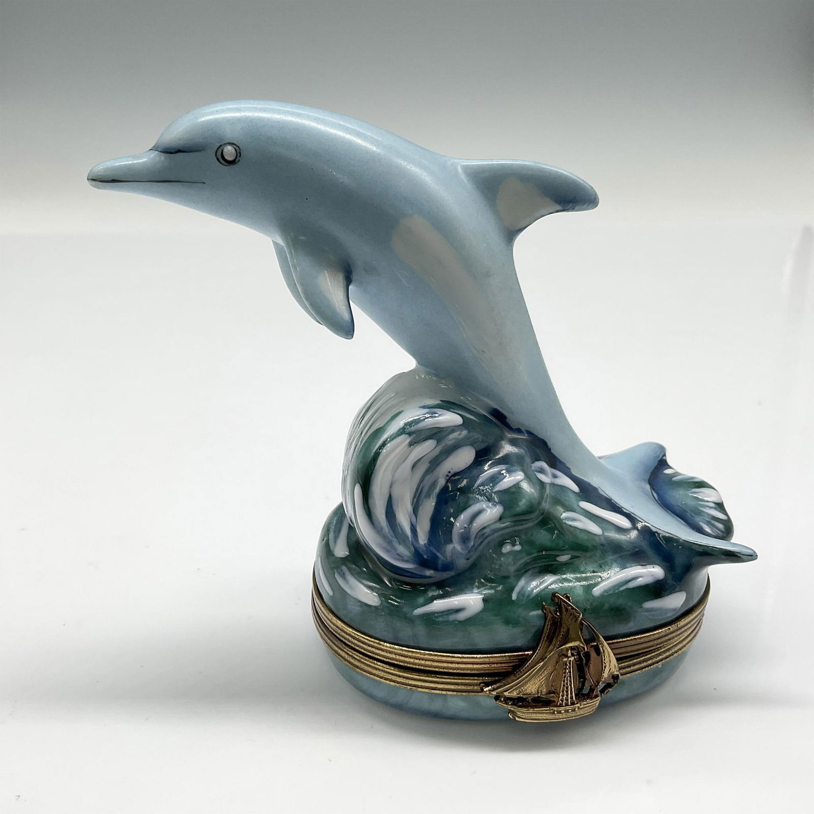 Dubarry Limoges Peint Main Porcelain Pill Box: Pale blue figure of a dolphin riding a wave. Metal banded, hinged lid with sailing boat as clasp closure. Lid opens to painted wave. Dubarry Limoges France backstamp. Issued: 20th century Dimensions: