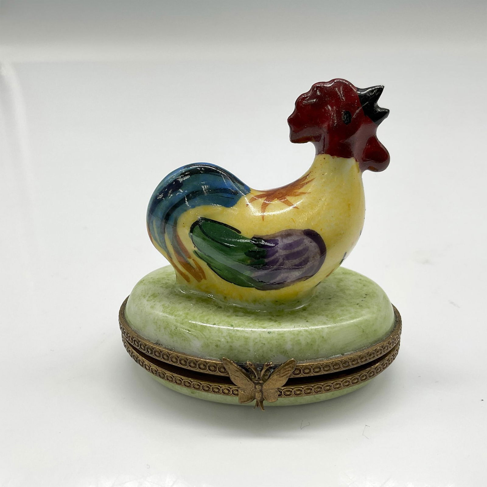 Limoges Porcelain Pill Box, Rooster: Colored in yellow, red, green and blue this colorful fellow sits on base of marbled green. Butterfly metal clasp closure opens to green leaf. Limoges France backstamp. Issued: 20th century Dimensions: