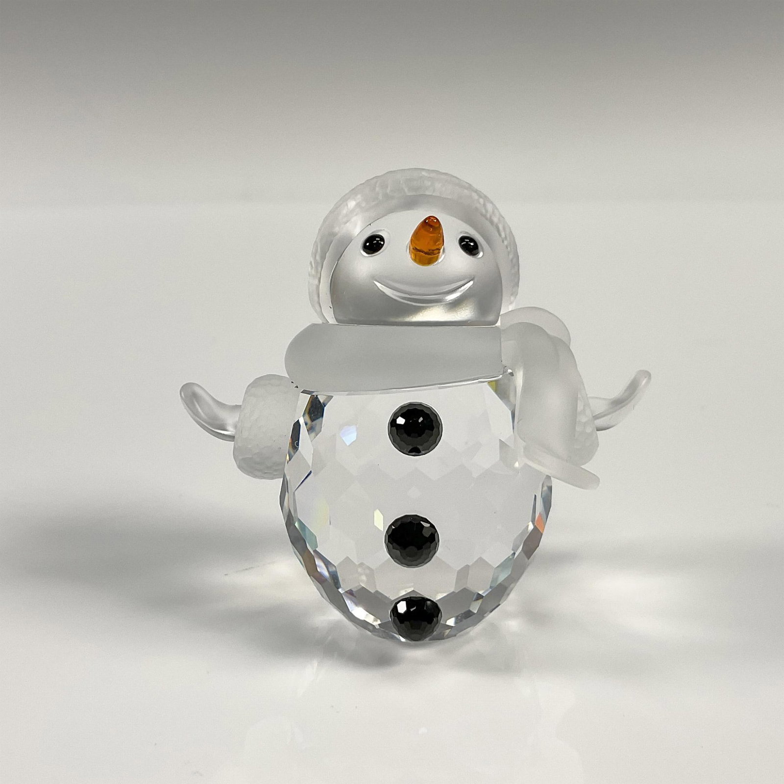 Swarovski Crystal Figurine, Snowman: Whimsical faceted crystal figurine modeled as a smiling snowman with his arms wide. Black buttons, orange nose. Satin crystal scarf, arms, and hat brim. Swarovski mark. 250229. This item has its origi