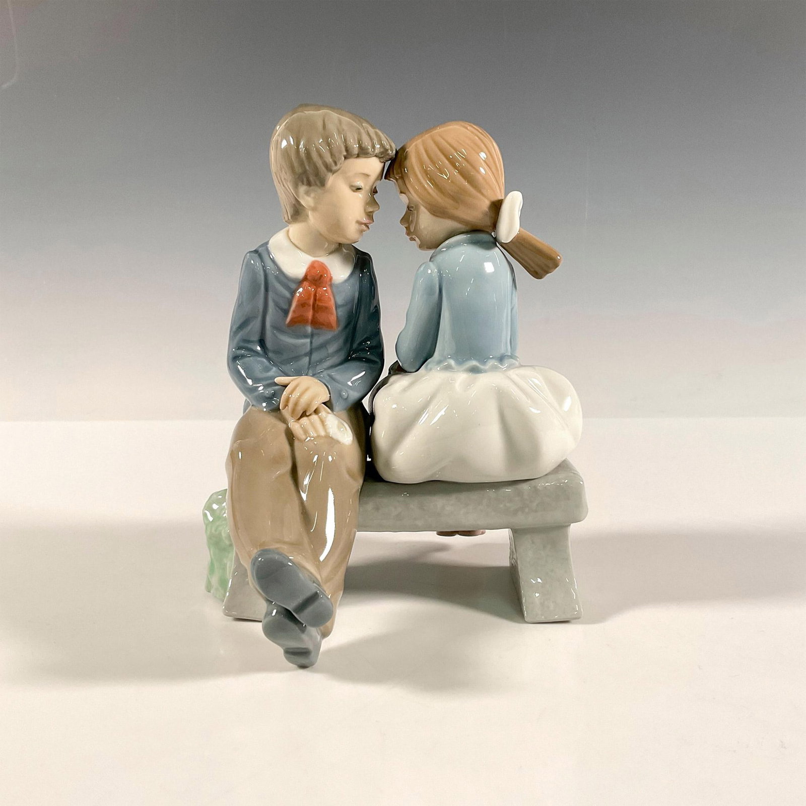 First Kiss 1136 - Nao By Lladro Porcelain Figurine: A boy and girl sitting on a bench about to begin the journey of love. Glossy finish. Nao by Lladro backstamp. Artist: Vicente Martinez Issued: 1990 Dimensions: 5.5"L x 5.5"W x 6.75"H Manufacturer: Nao